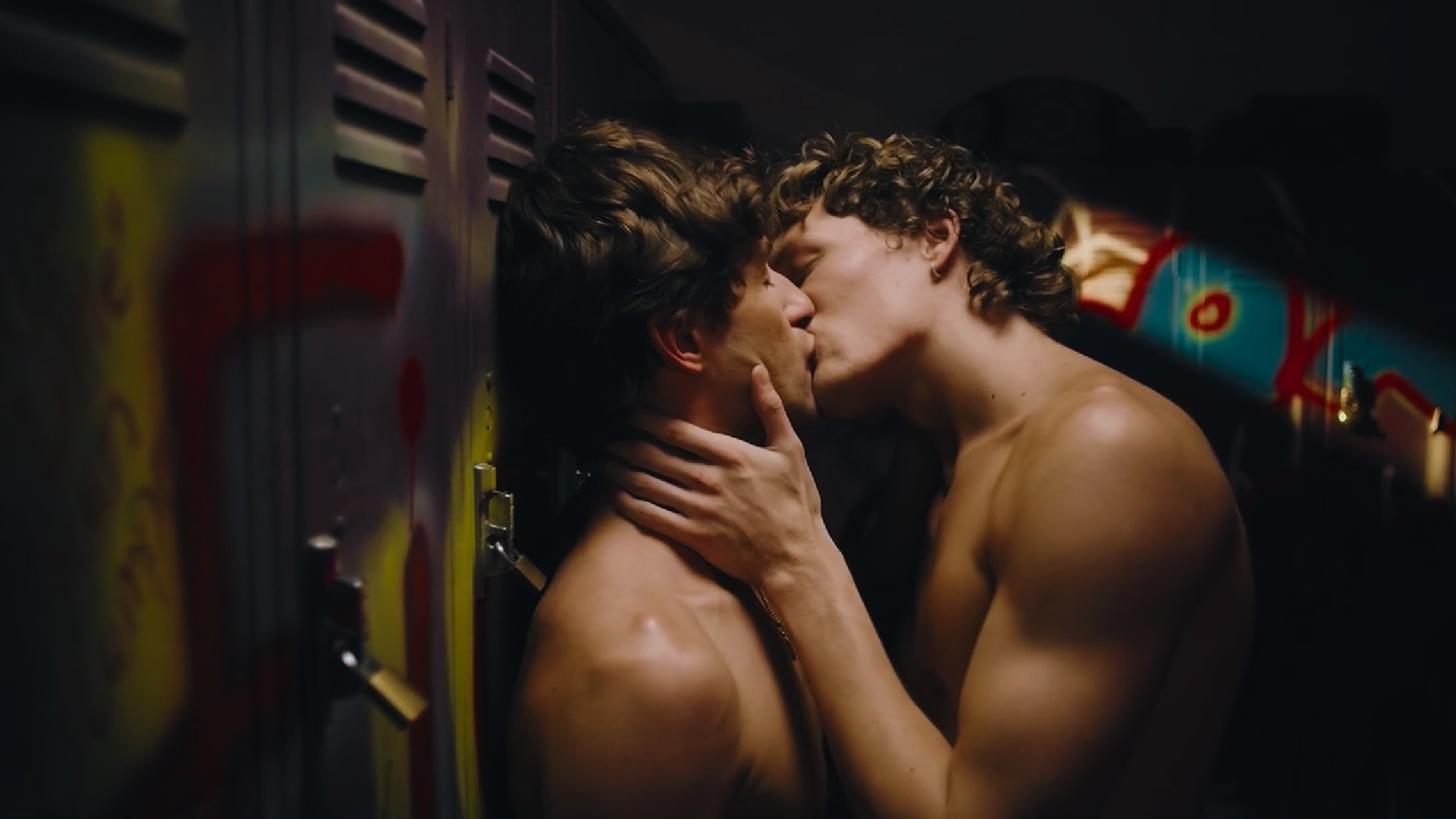 Two shirtless young men kissing in a locker room, with lockers in the background.