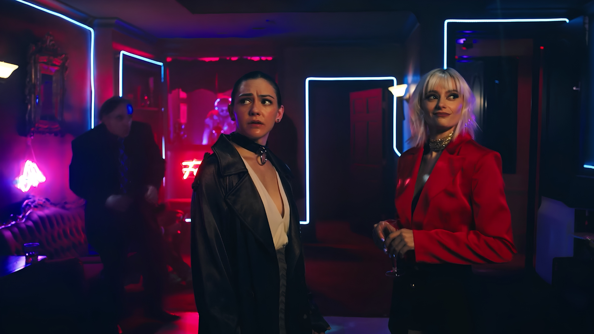Two women standing in a neon-lit bar, one with dark hair and leather jacket looking worried, the other with blonde hair and red blazer smiling, with a man in the background.