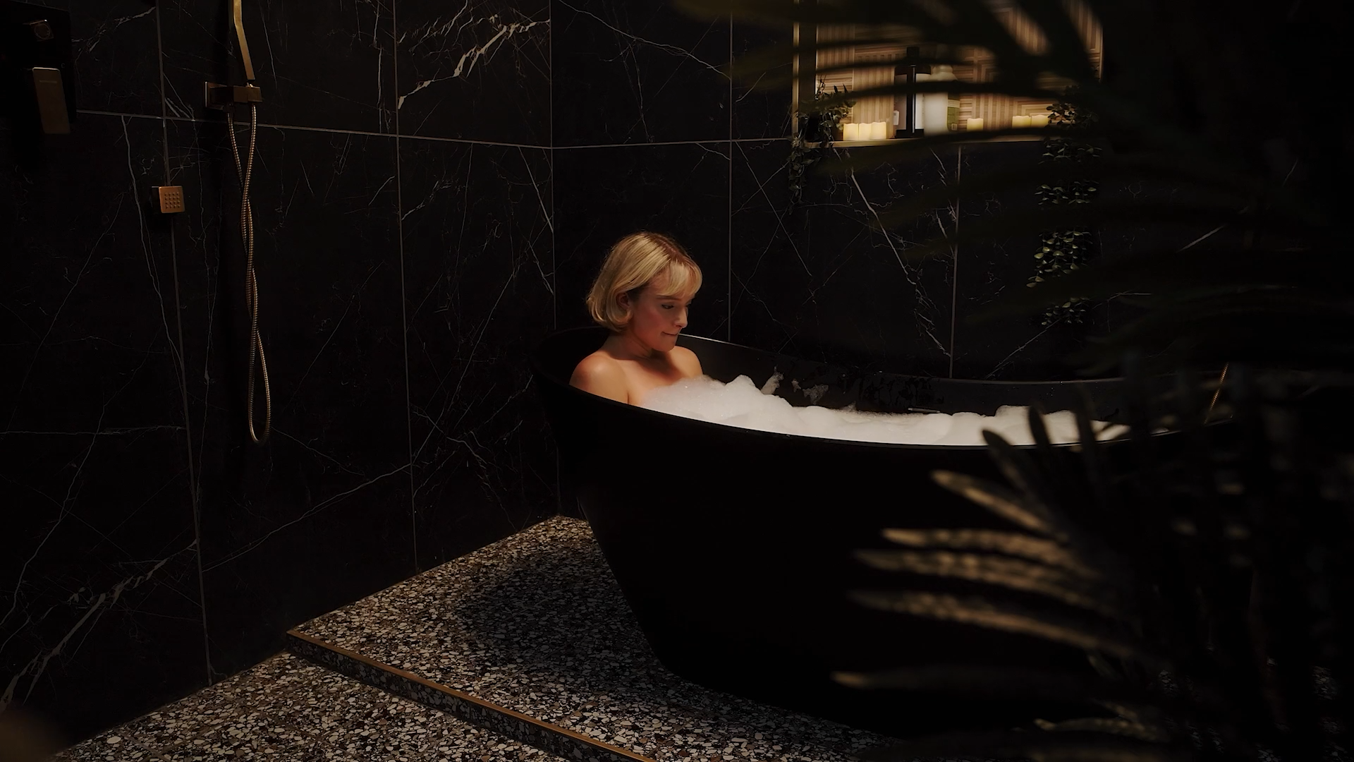 A woman with blonde hair relaxing in a black bathtub filled with bubbles in a dark, stylish bathroom with black marble tiles and a small window.