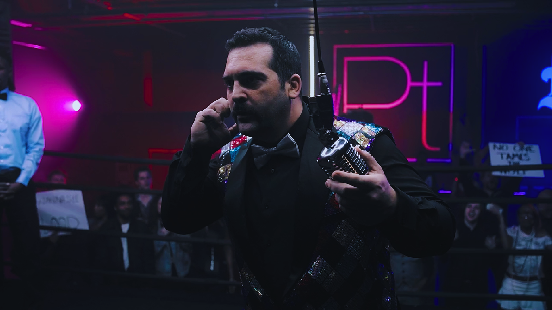 A man in a black sequined jacket with colored patches, holding a vintage microphone, talking on a phone inside a neon-lit boxing ring, with an audience in the background.