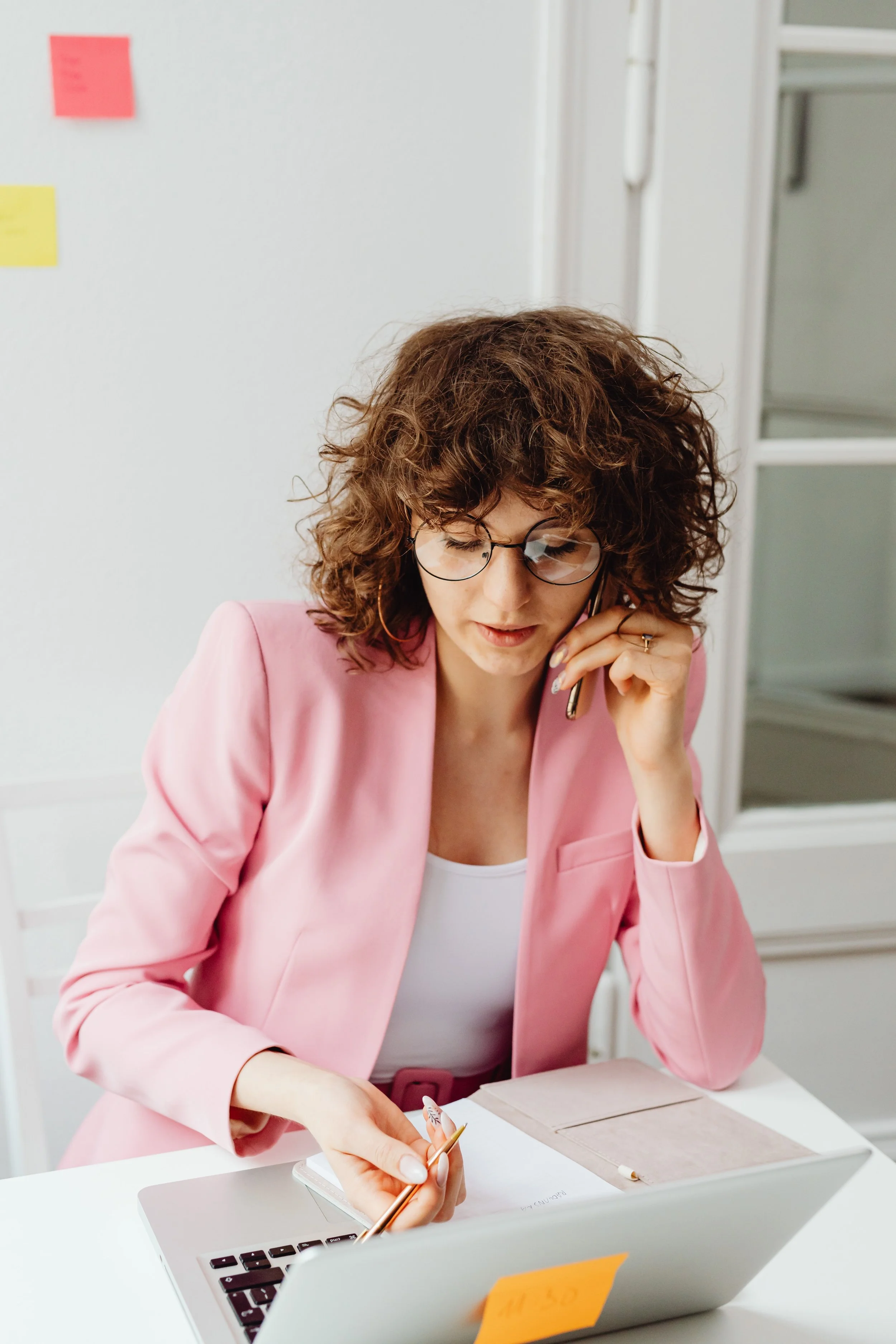 Woman with curly hair and glasses in a pink blazer talking on a cellphone at a desk with a laptop and notebook.