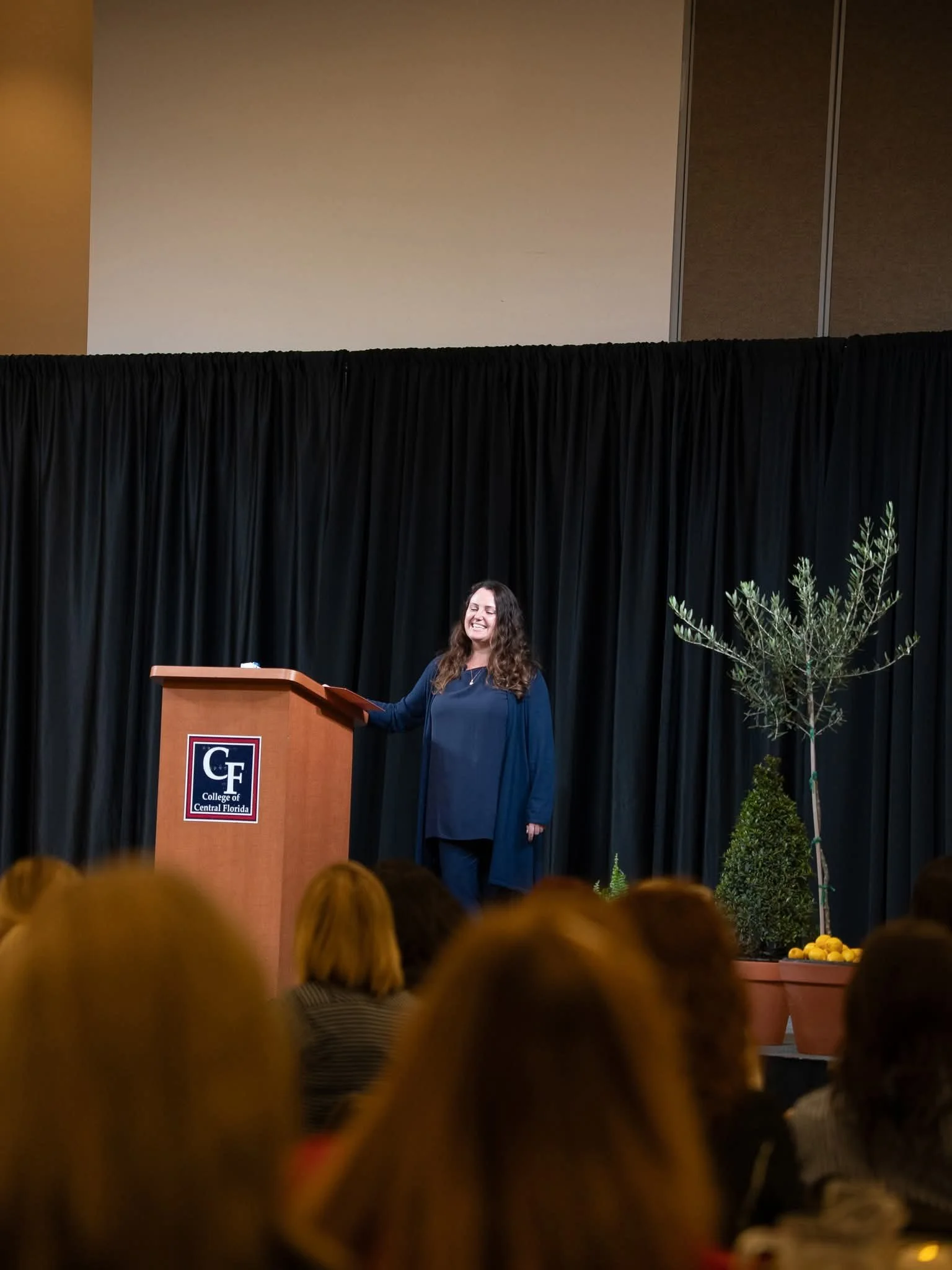A woman standing at a podium on a stage with a black curtain backdrop, smiling, with plants on her right side, in front of an audience.