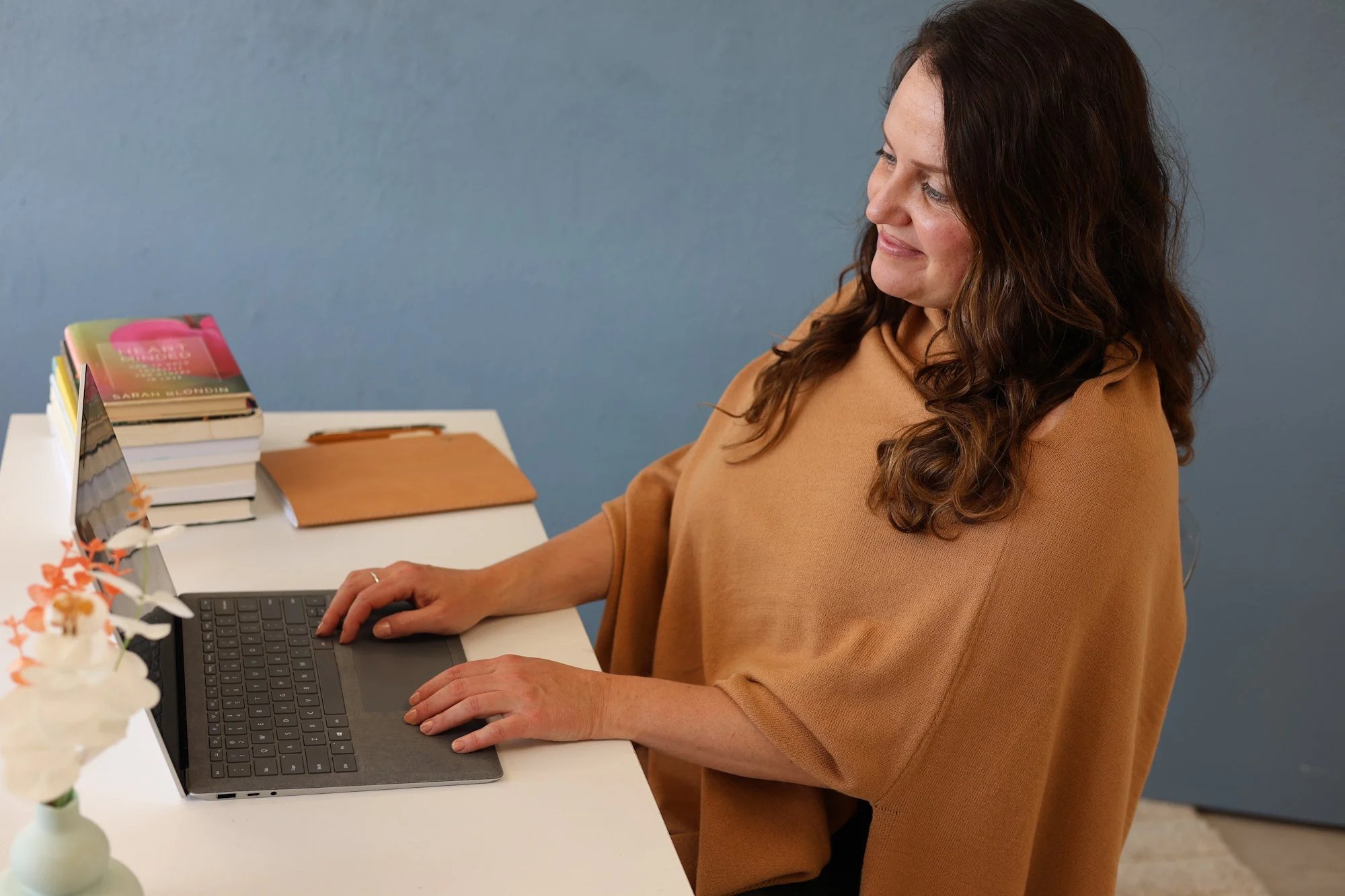 A woman with long, wavy brown hair wearing a camel-colored top sitting at a white desk, working on a laptop with a black keyboard. There are stacks of books, a brown folder, a small decorative plant, and a blue wall in the background.