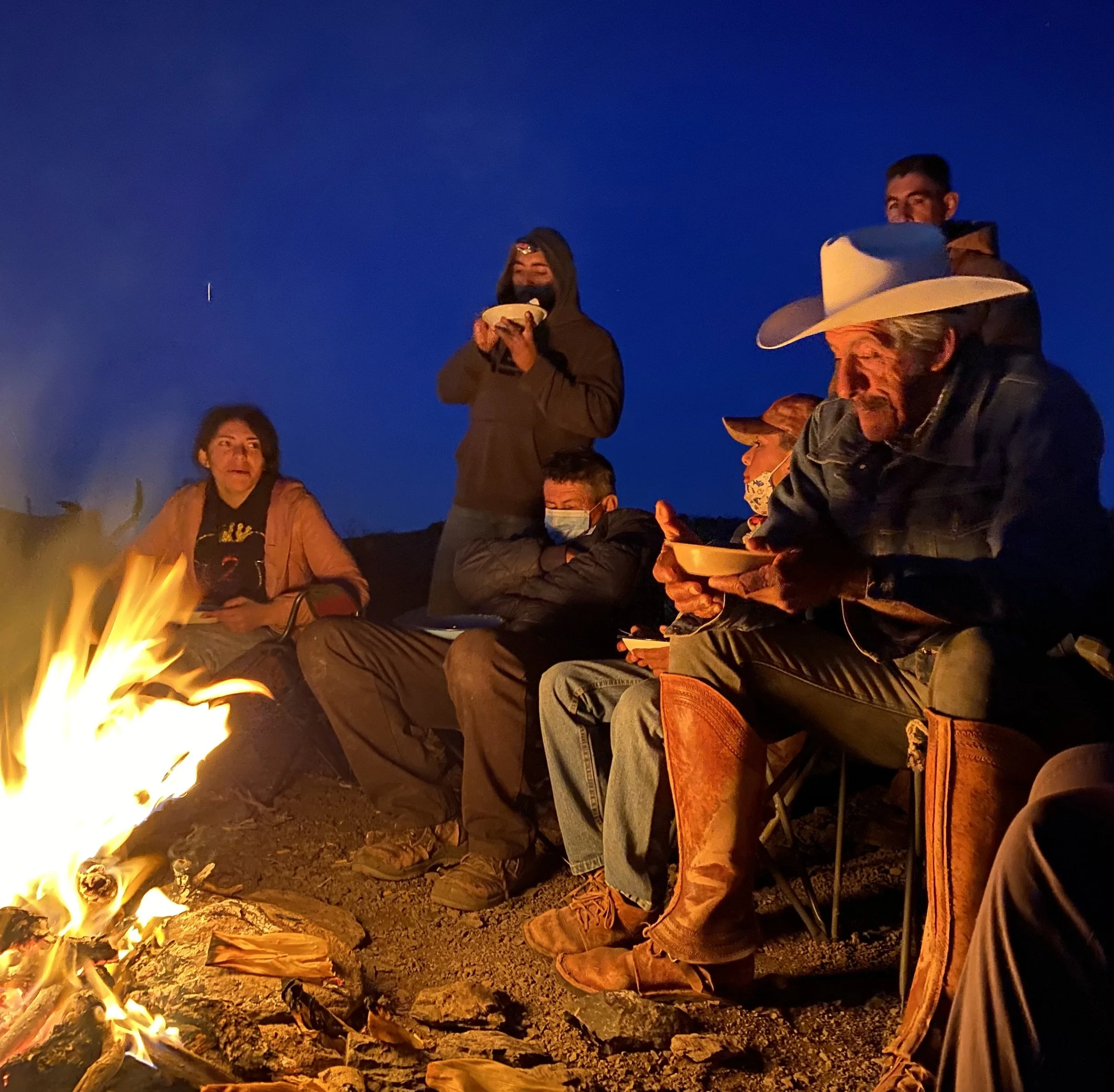 Group of people sitting around a campfire at night, some wearing masks, with clear skies overhead.