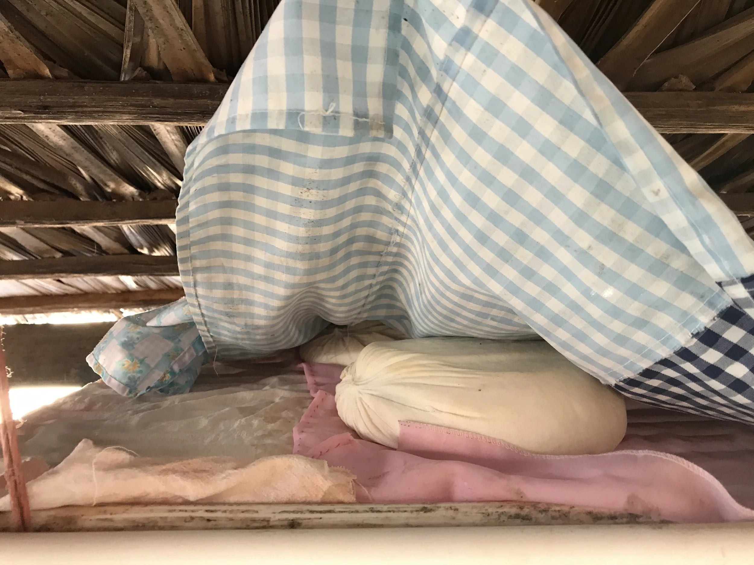 A bundle of fabric, including a blue and white striped cloth, resting on a pink cloth with a pillow or bundle of some sort underneath in an attic or storage space with exposed wooden beams.