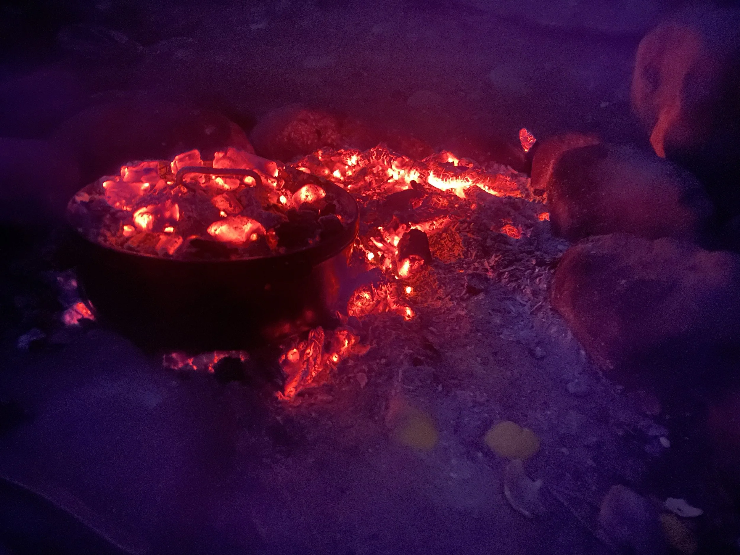 A black pot placed over glowing red hot embers and ashes, surrounded by rocks at night.