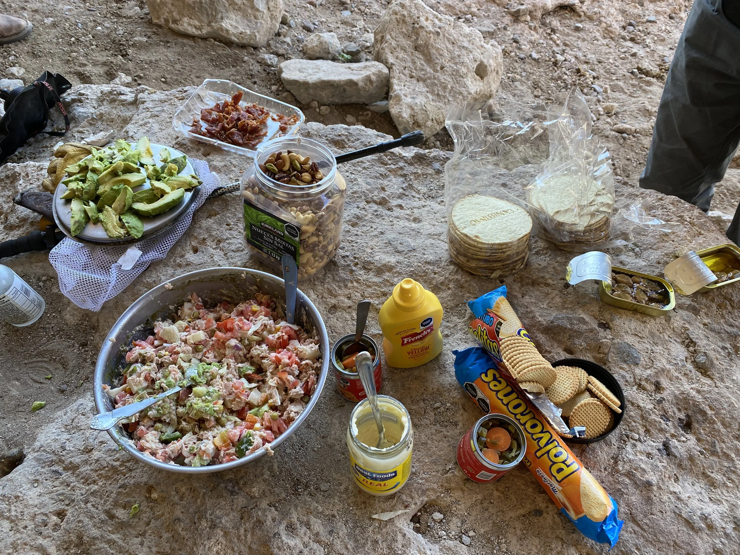 A messy outdoor picnic spread on a rocky surface includes a large bowl of salad, a plate of avocado slices, canned beans, packets of crackers, a jar of mustard, a jar of mayonnaise, and various other snacks and food items.