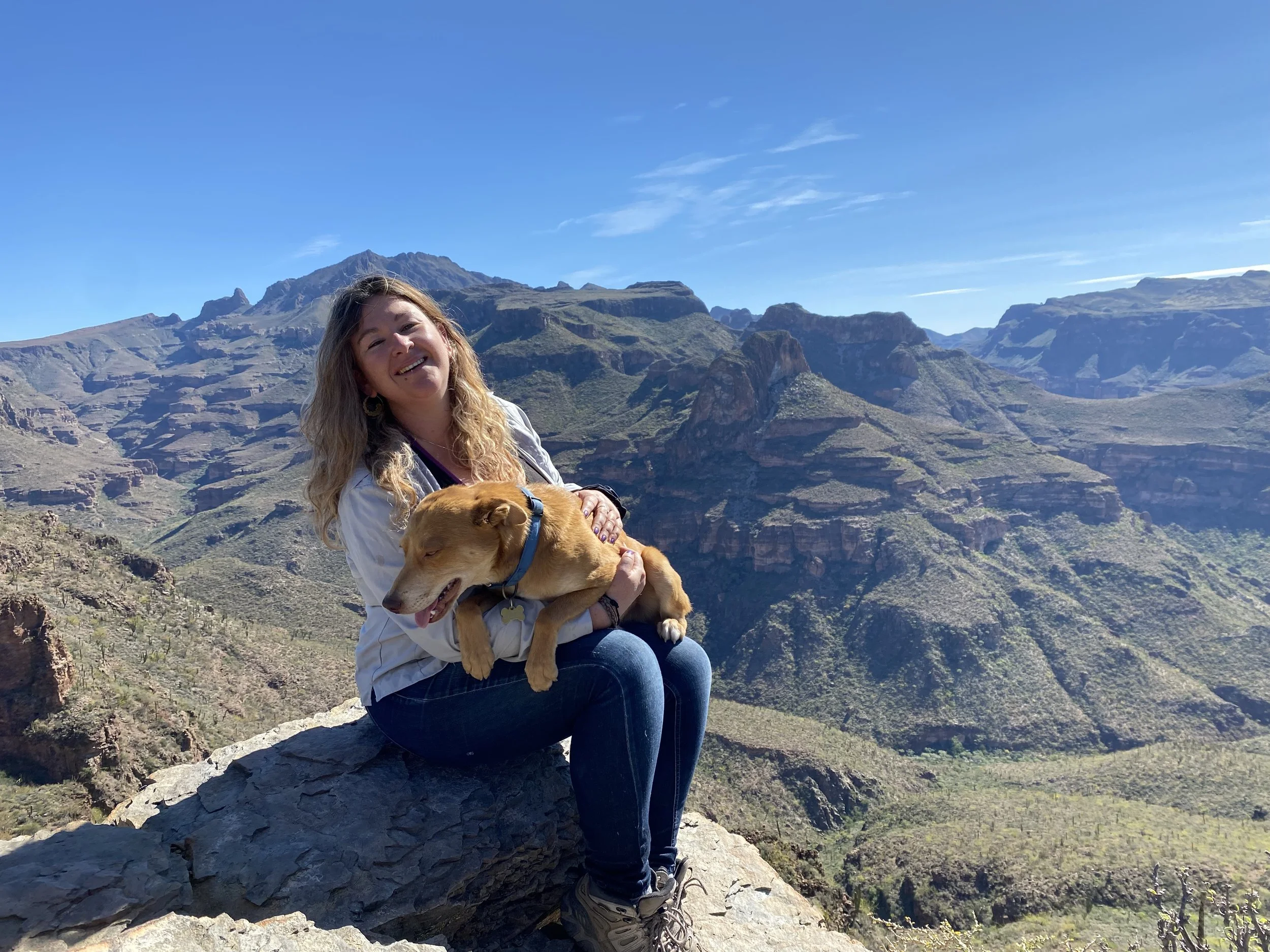 A woman is sitting on a rock in a mountainous landscape with a dog, smiling at the camera under a clear blue sky.