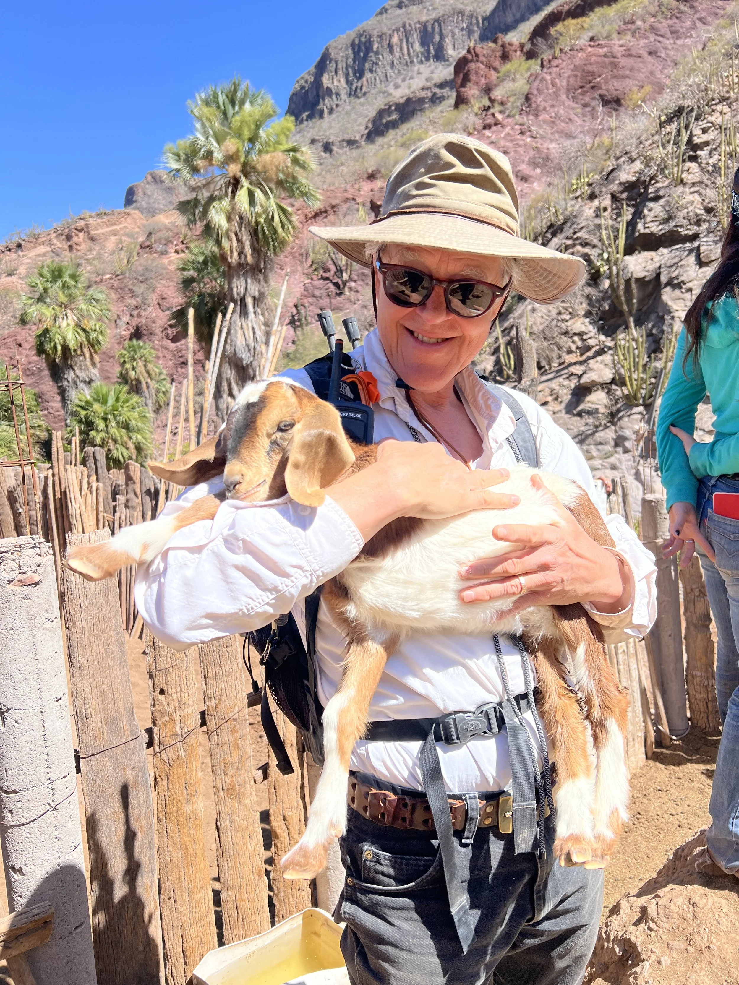 A smiling woman wearing a wide-brimmed hat, sunglasses, and outdoor clothing, holding a small goat in her arms, with desert mountains, cacti, and palm trees in the background.