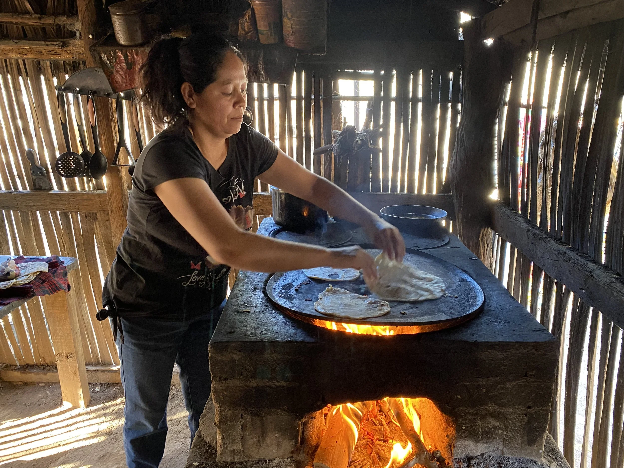 A woman cooking flatbread on a traditional outdoor stove with an open flame, inside a rustic wooden kitchen.