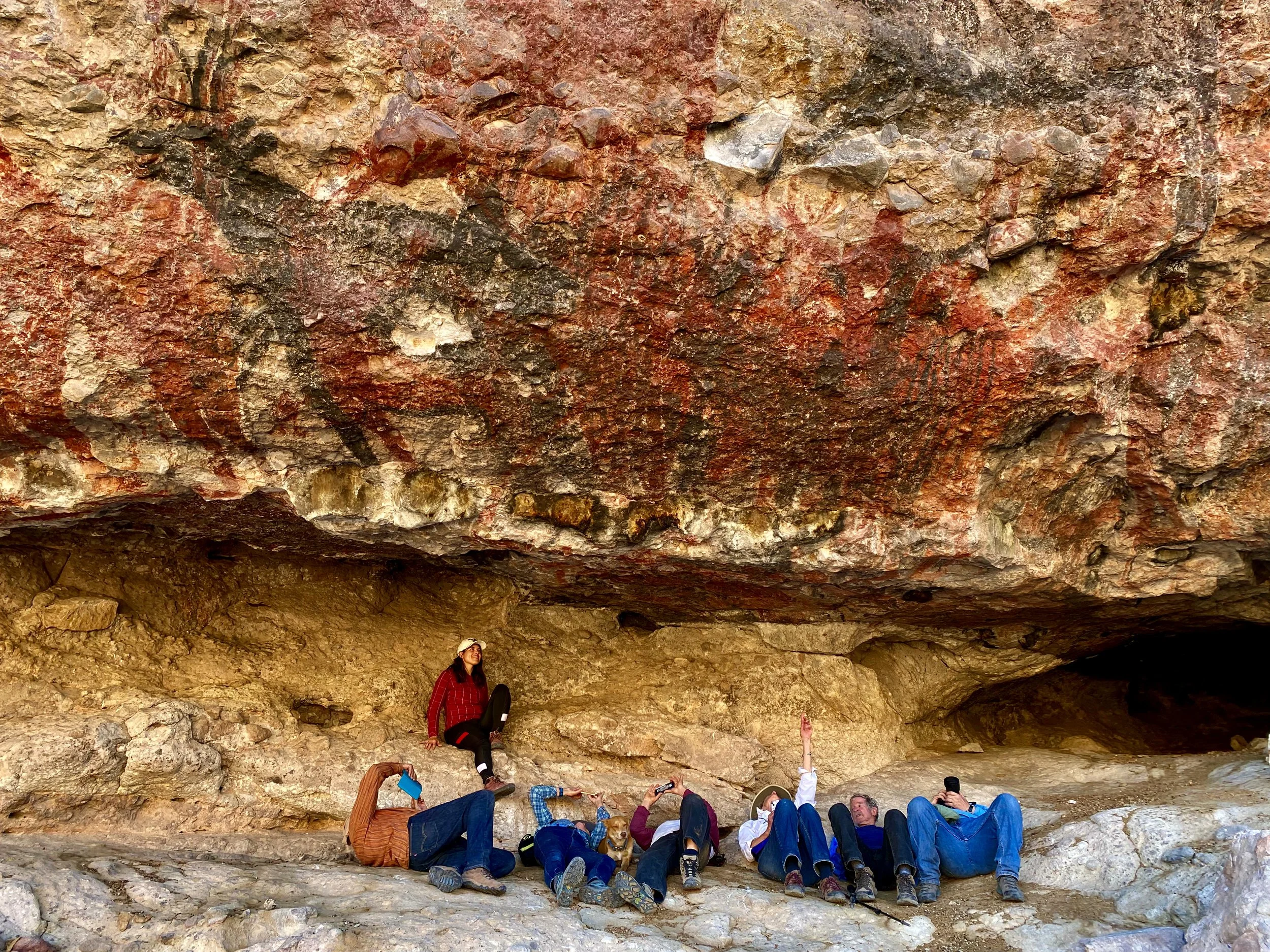 Group of people sitting and lying on rocky ground under a large overhanging rock formation, with one person standing and smiling nearby. The rock has a reddish-brown color and textured surface.