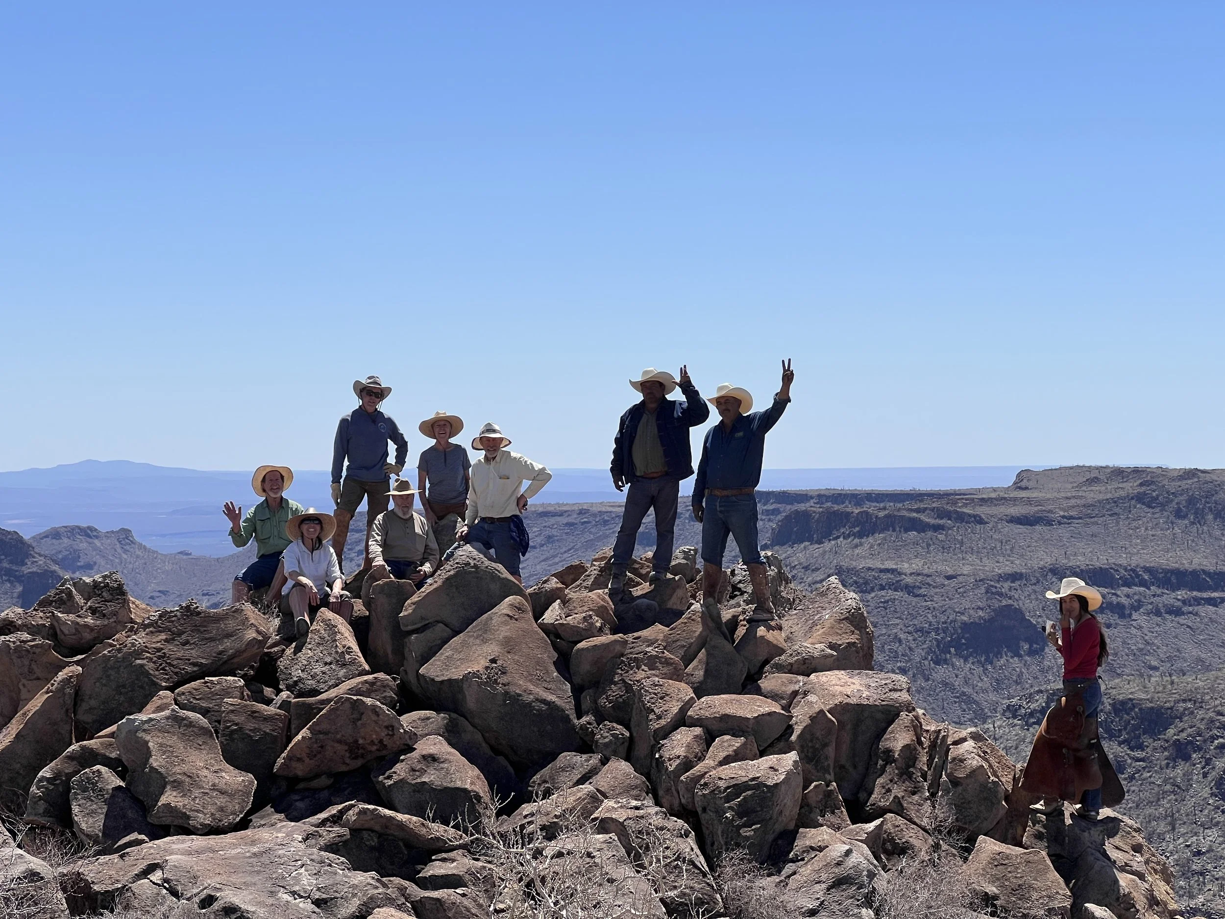 Group of people on rocky terrain in a desert, wearing cowboy hats and casual outdoor clothing, posing for a photo with some making peace signs, with a vast canyon landscape in the background under a clear blue sky.