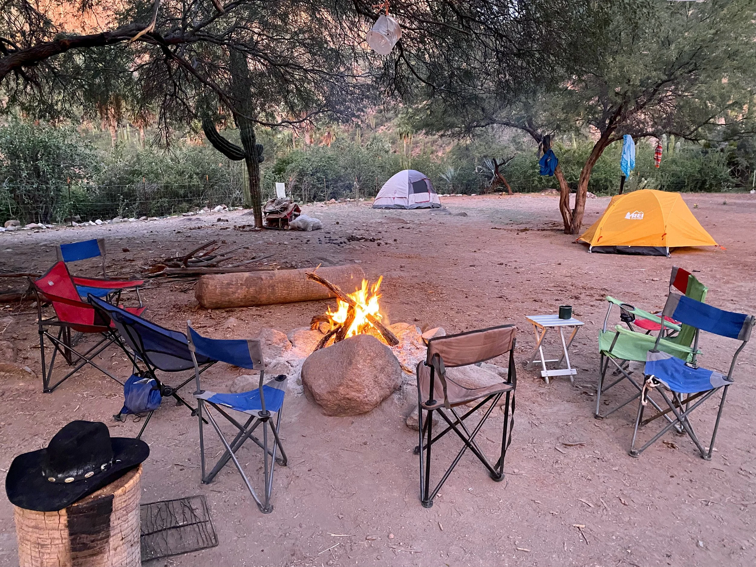 Camping scene with six camping chairs arranged around a campfire, two tents in the background, trees and bushes, and various camping gear.