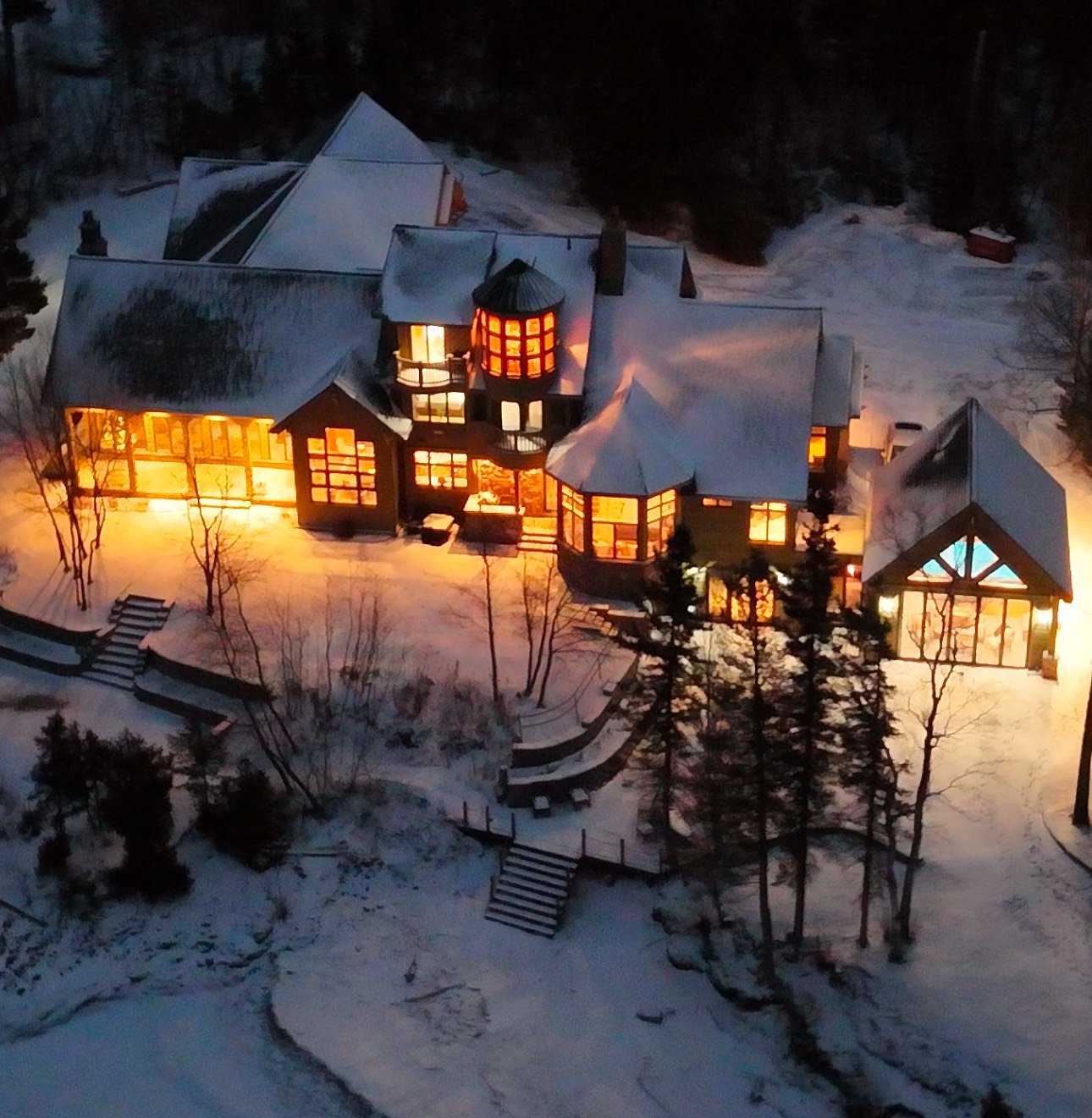 A large house illuminated from inside at night in a snowy landscape, with multiple sections, steep roofs, and a central tower with red window panes. The house is surrounded by snow-covered trees and steps leading down to a frozen or snow-covered grou