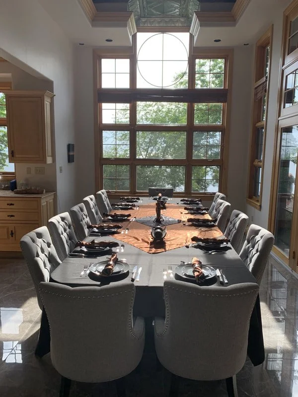A dining room with a long rectangular table set for a meal, with twelve upholstered chairs, twelve plates, silverware, and rolled napkins. Large windows with wooden framing and a view of greenery are in the background, allowing natural light into the