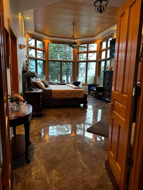 A bedroom with a large curved window wall overlooking trees, featuring a bed with pillows, a dark dresser, a small round side table with flowers, and a couple of chairs near the window, with a wood-paneled ceiling and marble floor.