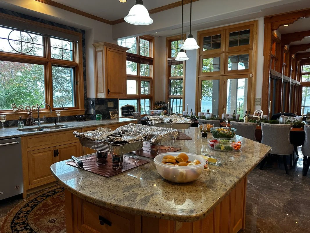 A spacious kitchen with wooden cabinets and large windows overlooking a lake. The kitchen island is set up for a meal or gathering, with foil-covered dishes, salads, and bread on the countertops. Dining chairs are arranged around a table in the backg