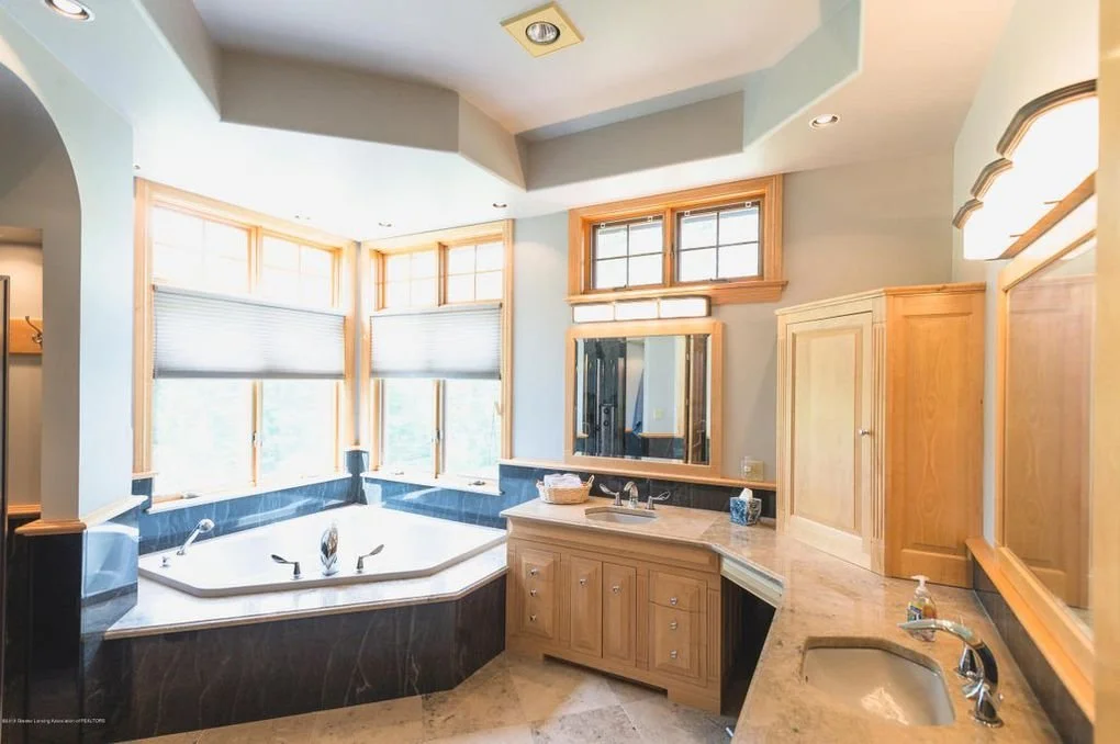 Bright bathroom with a corner soaking tub, multiple windows with wooden frames, a large mirror above the vanity, light wood cabinetry, and beige tiled countertops.