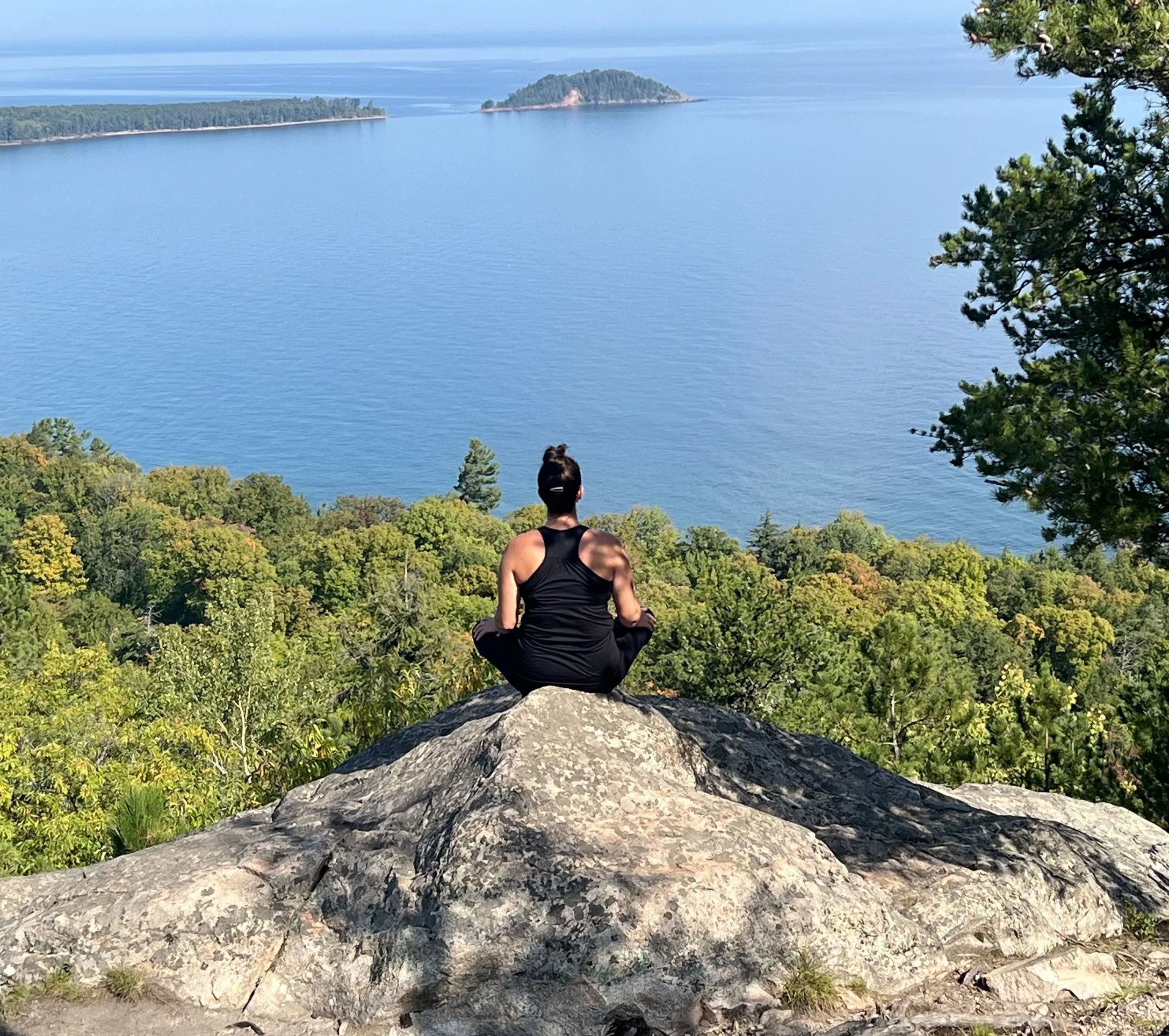 A person in black athletic clothing sitting cross-legged on a large rock, facing a body of water with trees in the foreground and islands in the background.