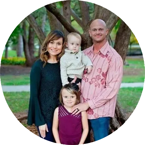 Family portrait of four standing outdoors under a large tree, with a woman, a man, and two young girls, smiling at the camera.