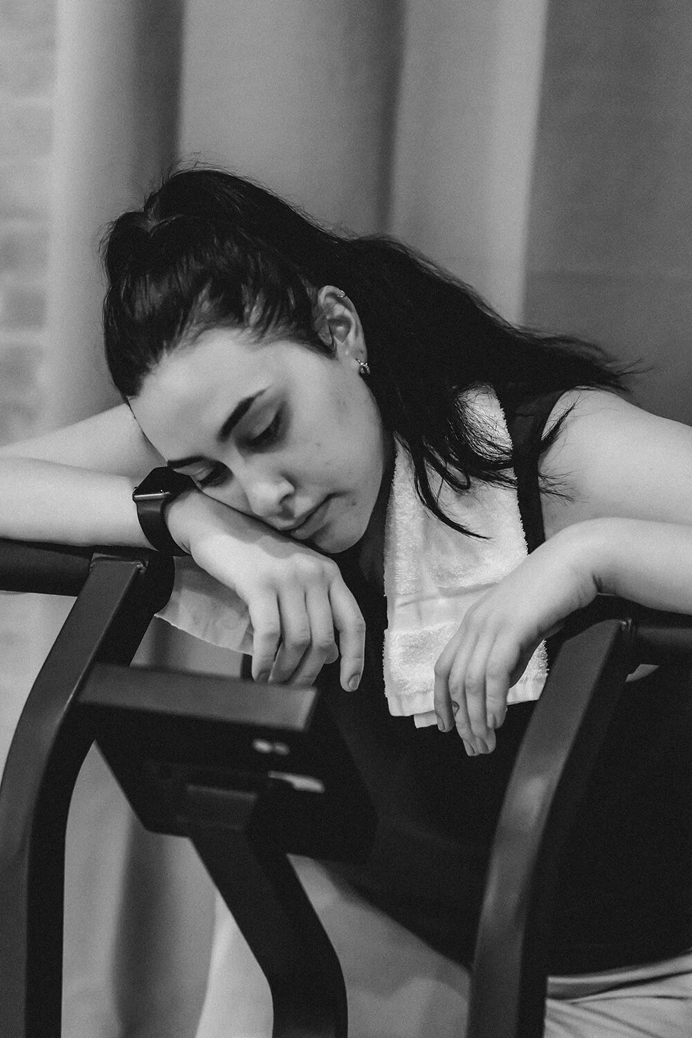 A young woman with dark hair resting her head on her arm on a chair, appearing tired or sad, in black and white.