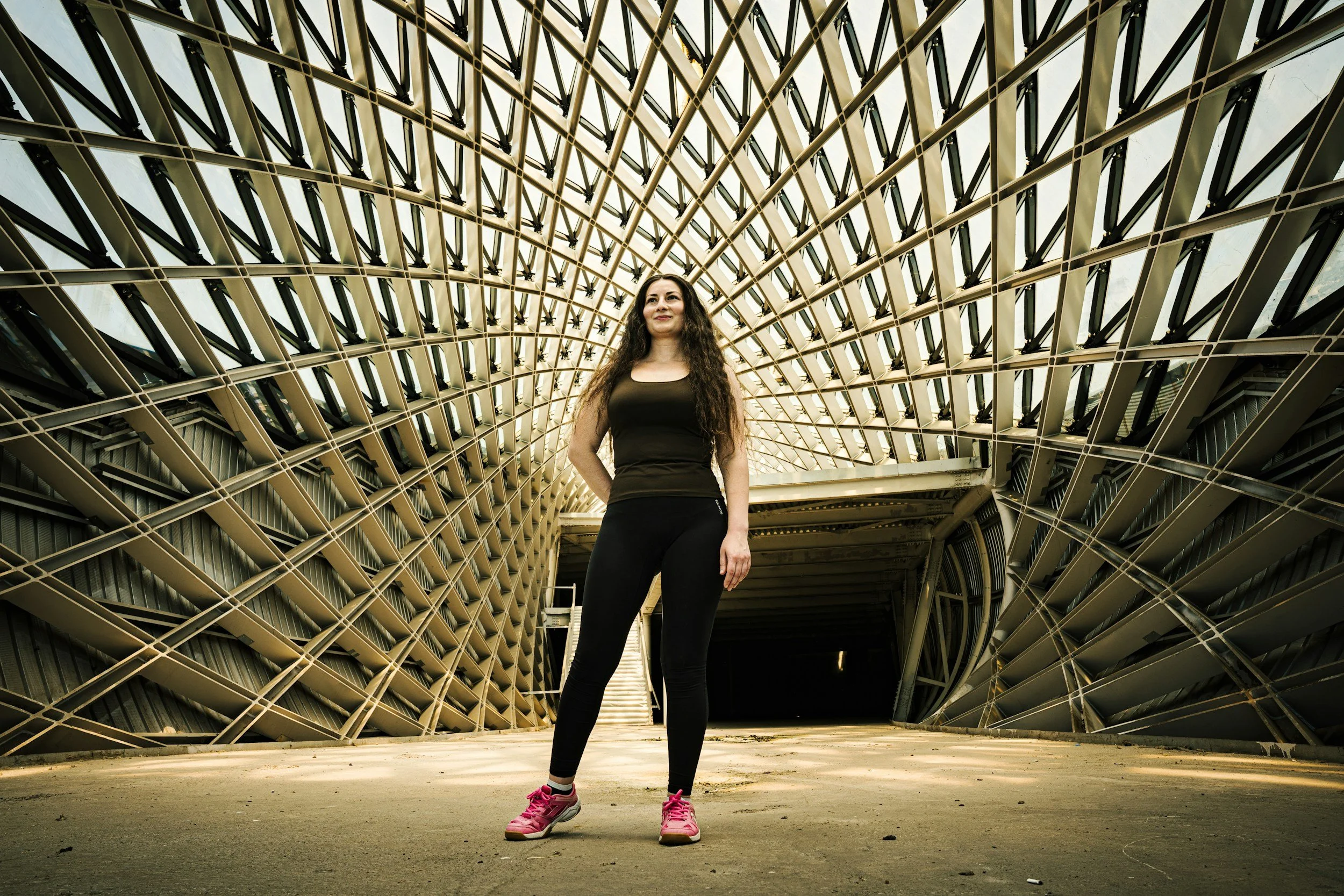 A woman in black athletic clothing and pink shoes standing outdoors in front of an architectural structure with a lattice design.