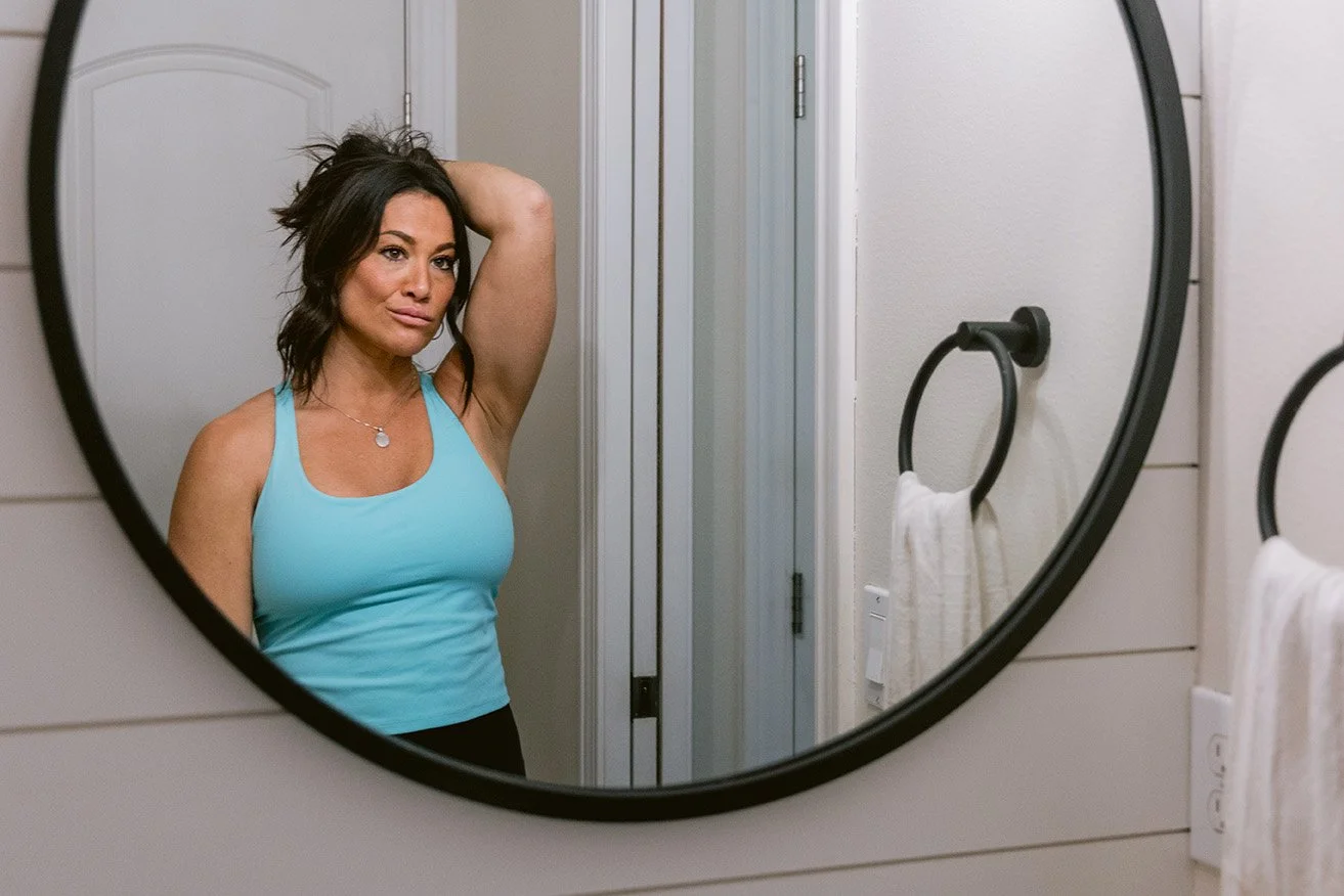 A woman with dark hair wearing a blue tank top taking a selfie in a bathroom mirror.