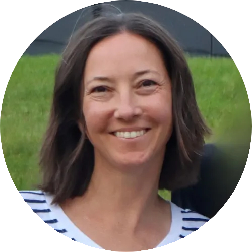 A woman with shoulder-length brown hair smiling outdoors with a grassy field and blurred background.