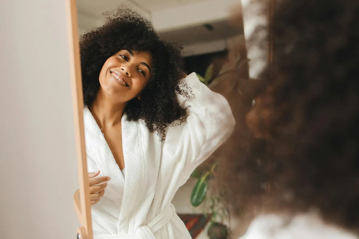 Woman with curly hair smiling and looking into a mirror, wearing a white robe.