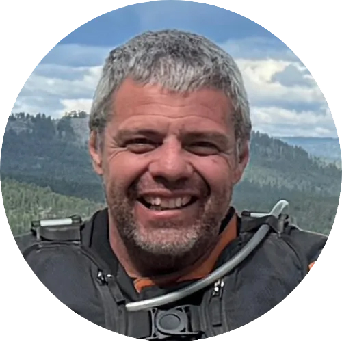 A smiling middle-aged man with short gray hair, a beard, and a black jacket outdoors with mountains and a cloudy sky in the background.