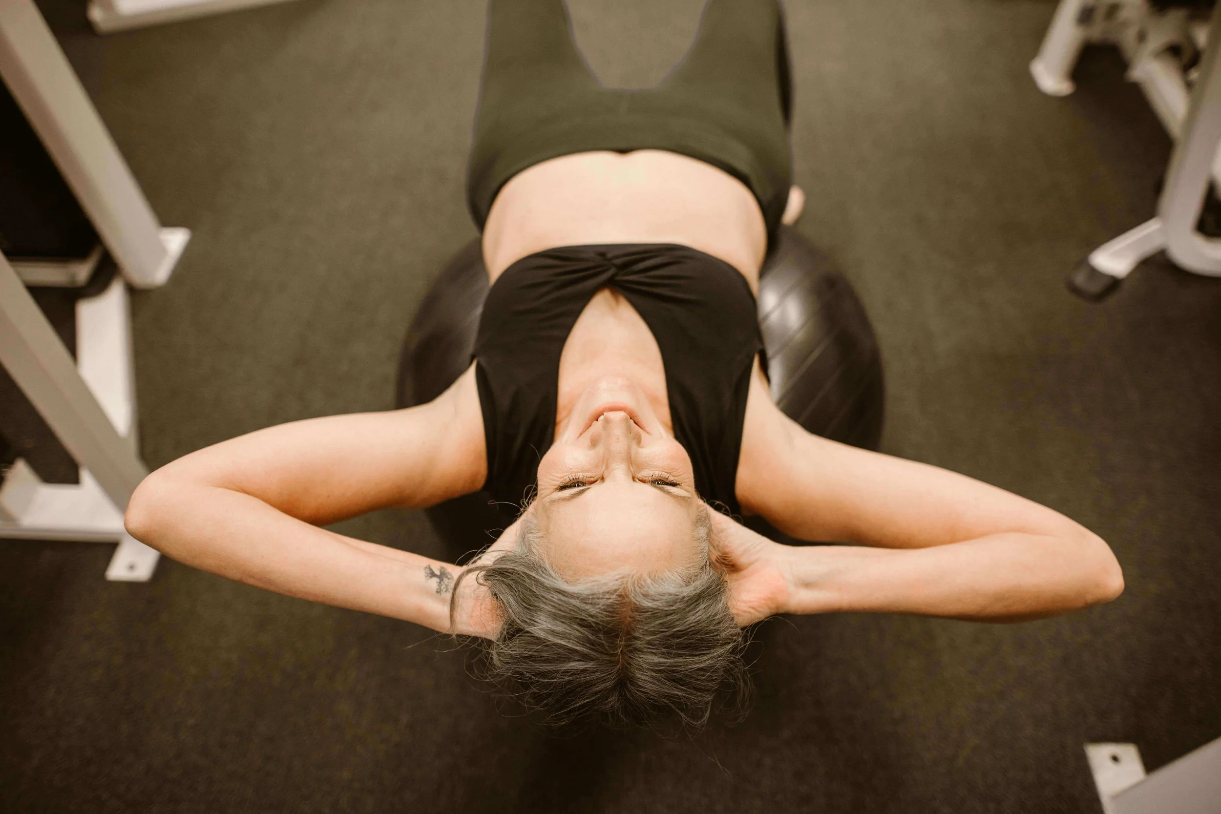 A woman lying on her back on a black exercise ball, with her hands behind her head and eyes closed.
