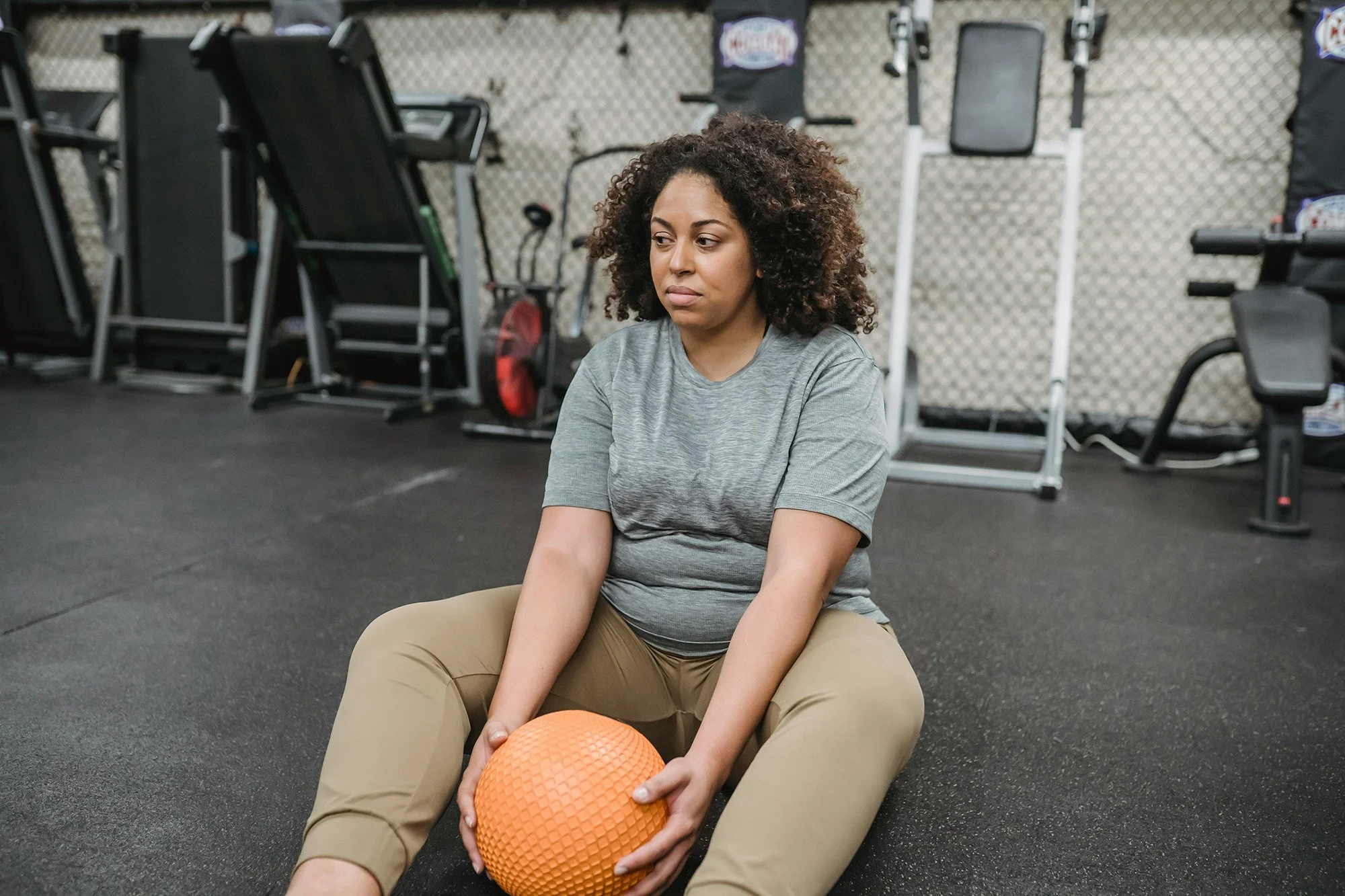 A young woman with curly hair sitting on the gym floor, holding an orange textured exercise ball between her legs, in a gym with workout equipment in the background.