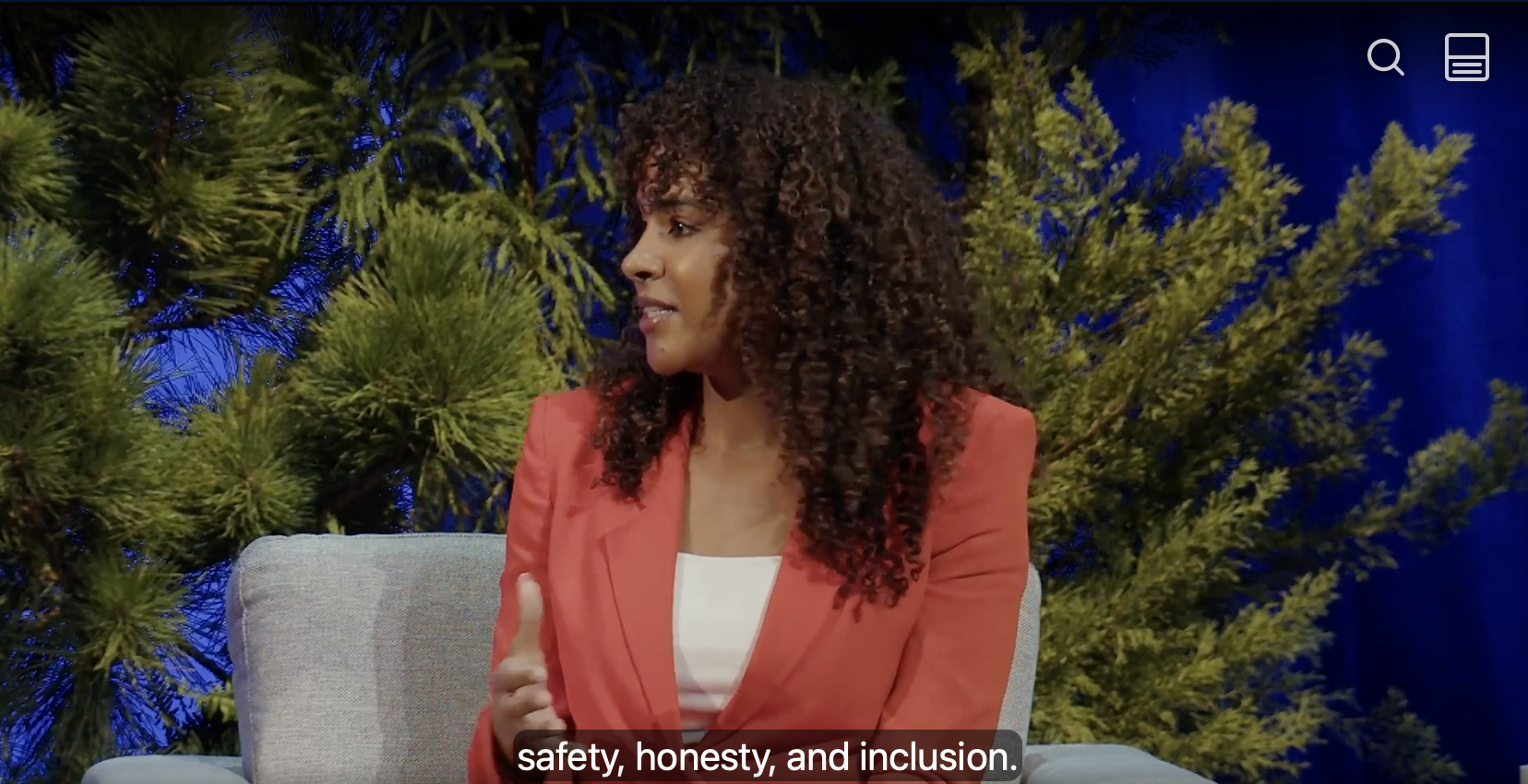 A woman with curly hair wearing an orange blazer and white top, sitting on a beige chair during a talk show or interview, with green pine tree branches in the background.