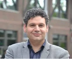A man with curly dark hair and a light complexion, wearing a gray blazer over a dark shirt, standing outdoors in front of a building and trees.