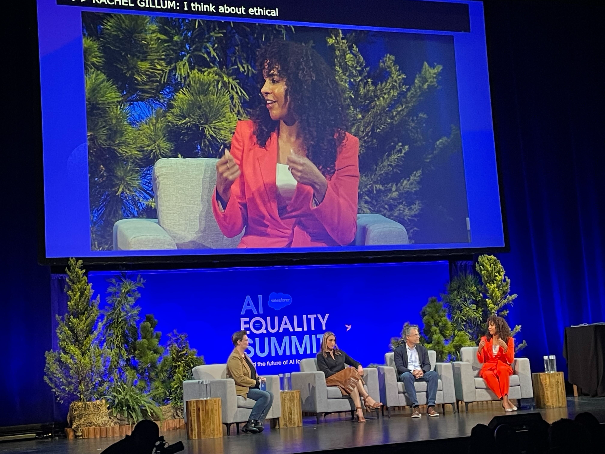 A woman in a red blazer speaking on stage at the AI Equality Summit, with a large screen behind her showing her close-up. Four other people are seated on stage with greenery and trees as the backdrop.