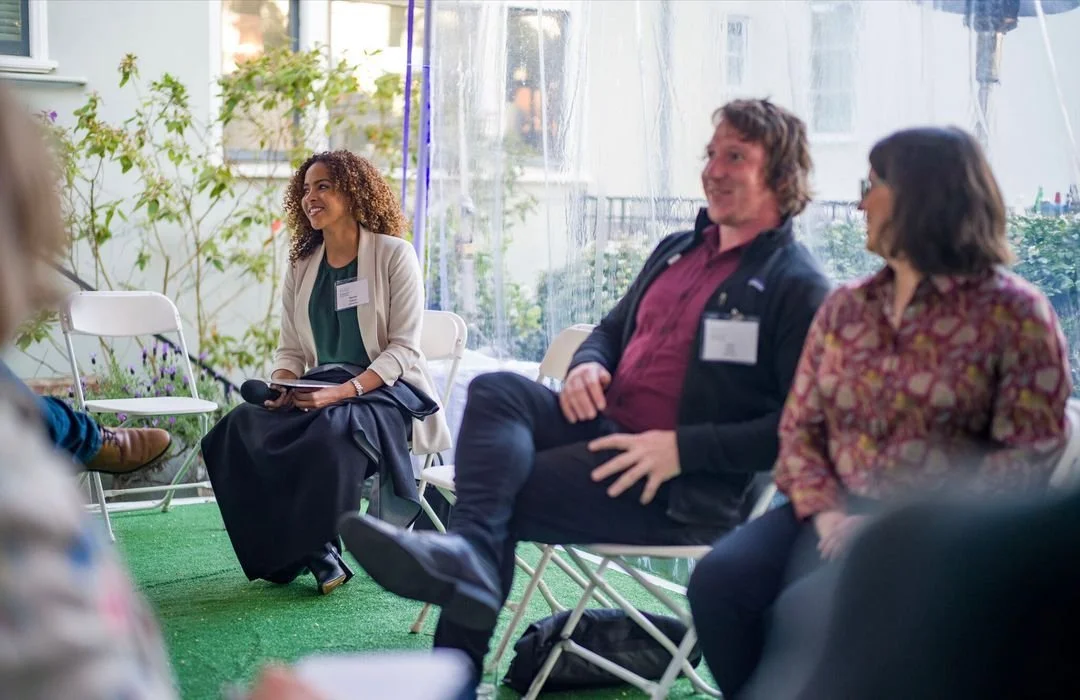 A group of people sitting in a semi-circle engaging in a discussion at an outdoor event, with plants and buildings in the background.