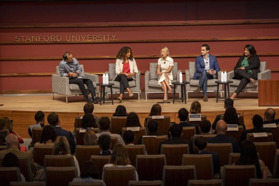 Panel discussion at Stanford University with five diverse speakers on stage, seated in gray armchairs with small tables holding water bottles, facing an audience in a large auditorium.