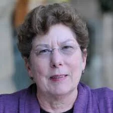 A portrait of an older woman with short, curly hair, glasses, and wearing a purple top, outdoors with a blurred background.