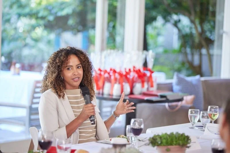 A woman with curly hair speaking into a microphone at a table during a formal event or meeting in a bright room with large windows and views of greenery outside.