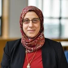 A woman wearing glasses and a patterned headscarf, smiling, indoors with a blurred window in the background.