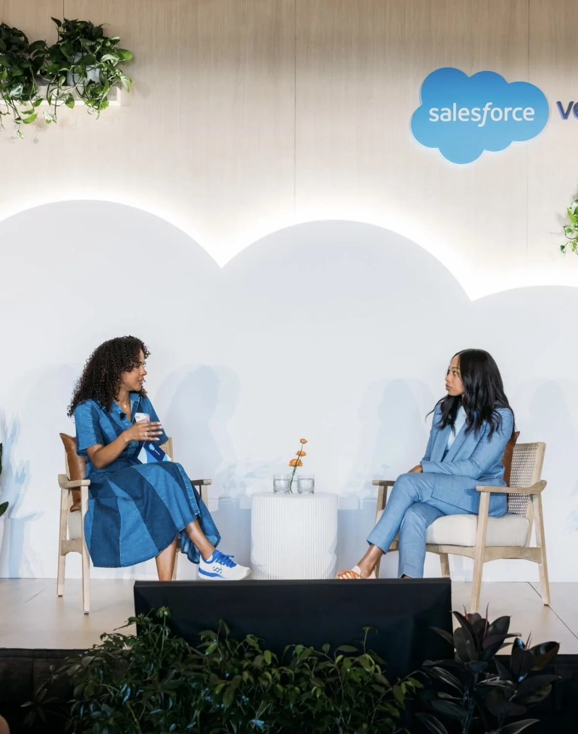 Two women (one is the famous American track star, Allyson Felix) sitting and talking on stage with a Salesforce logo backdrop, plants in foreground, and a small white table with flowers between them.