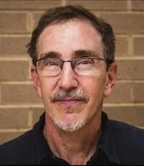A middle-aged man with glasses and facial hair, standing in front of a brick wall.