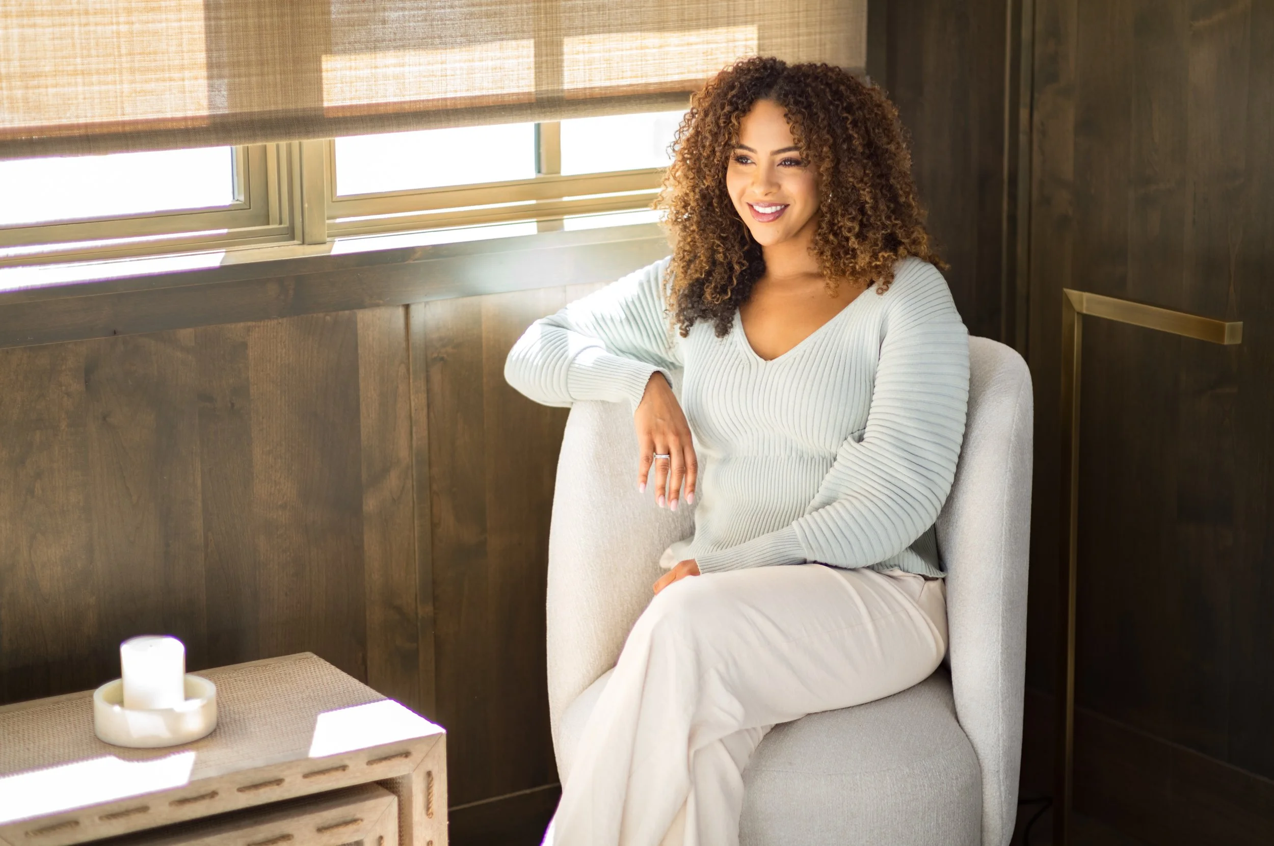 A woman with curly hair smiling while sitting on a beige chair near a window with blinds.