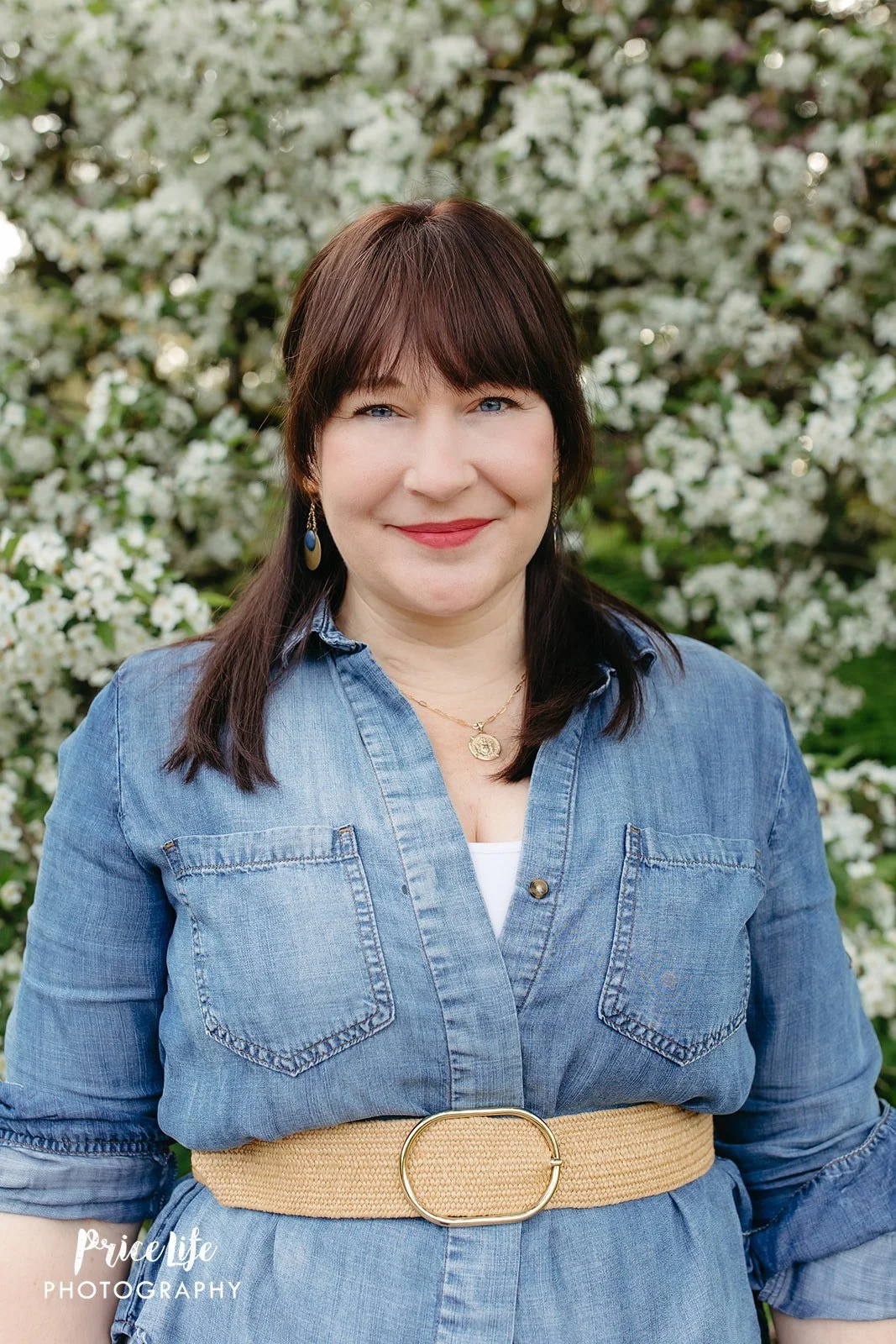 A woman with dark brown hair, blue eyes, and light skin standing outdoors in front of a flowering tree, wearing a denim dress with a beige belt, gold necklace, and earrings.