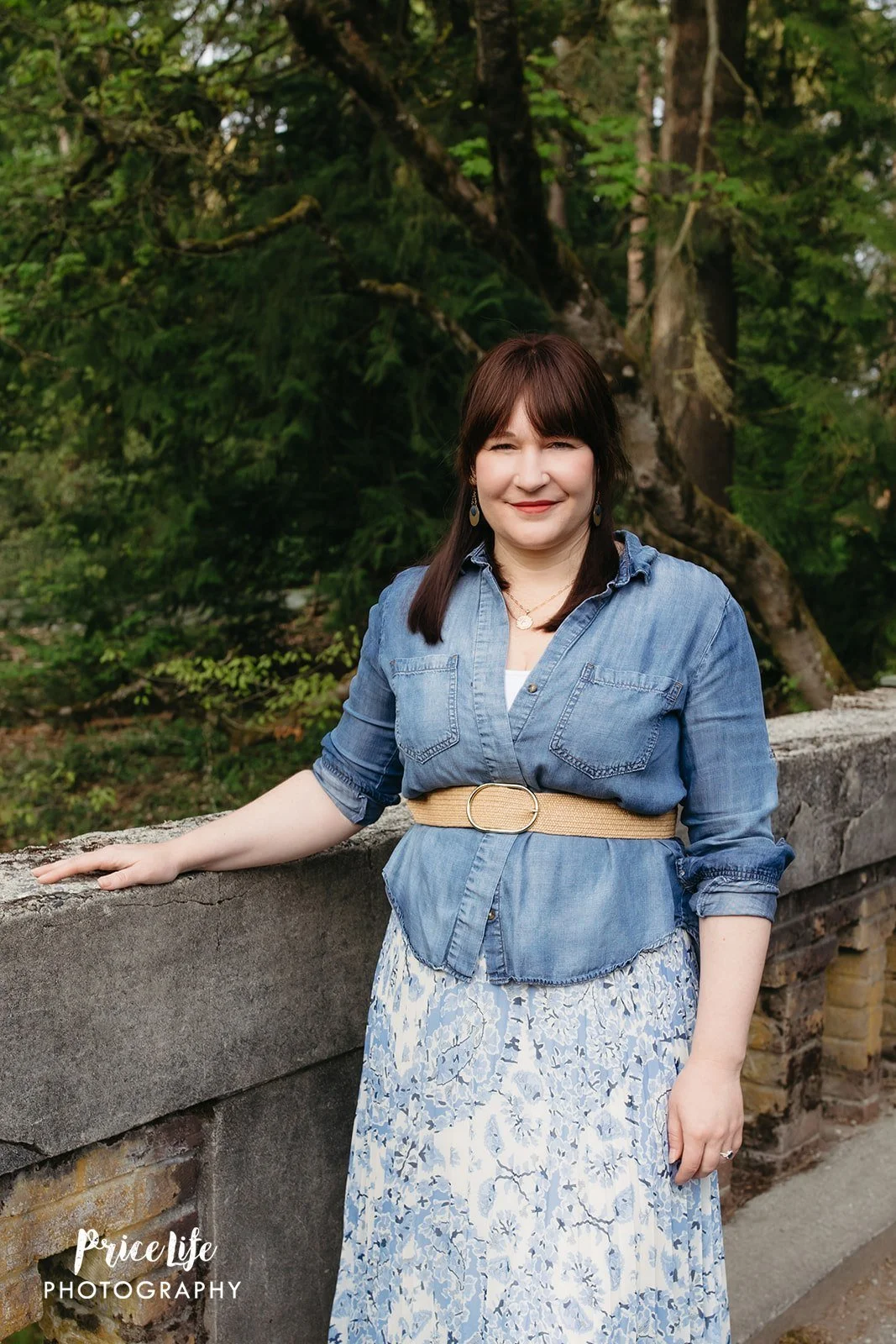 A woman with dark brown hair, wearing a denim shirt belted at the waist, and a white and blue floral skirt, standing outdoors by a stone wall with green foliage and trees in the background.