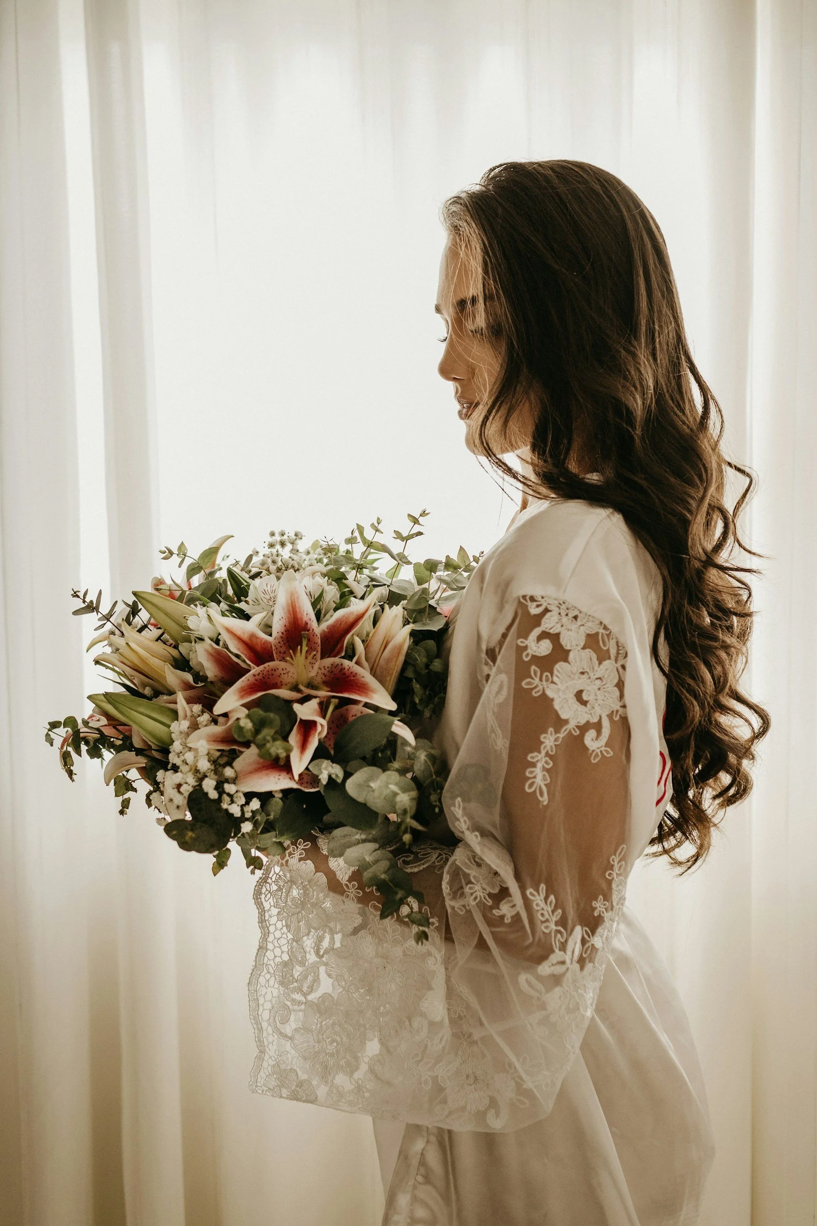 A woman in a white dress holding a bouquet of pink and white lilies and greenery, standing by a sheer curtain.