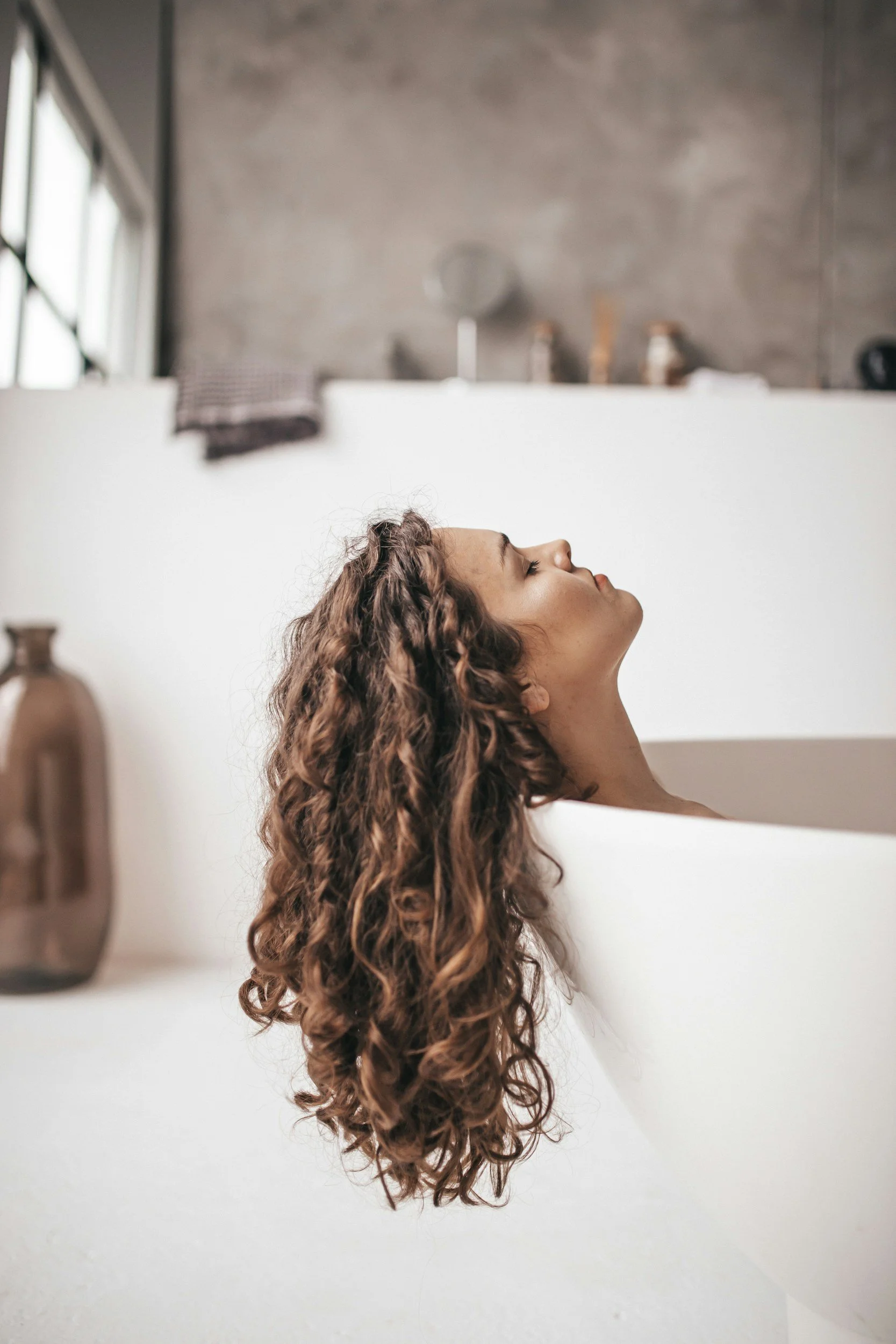 A woman with curly brown hair relaxing in a white bathtub in a modern, minimalist bathroom.