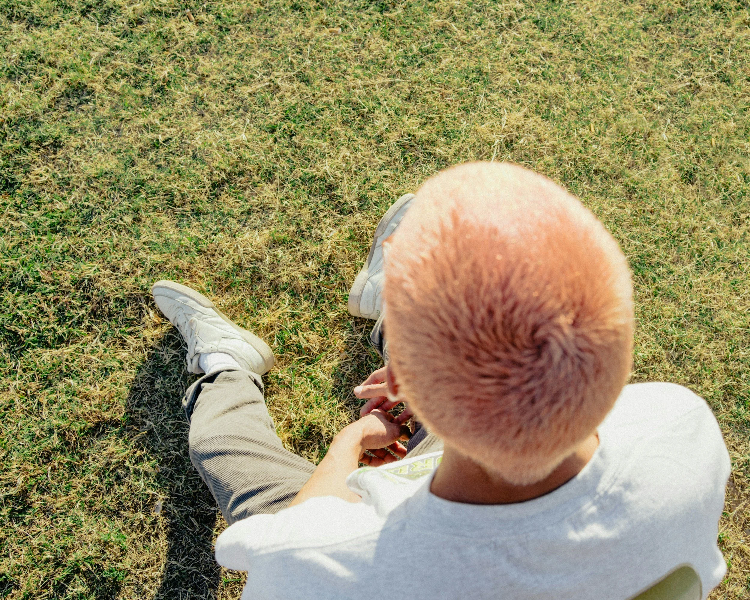 A person with short, dyed orange hair sitting on grass, wearing white sneakers, khaki pants, and a white T-shirt, with a shadow cast on the grass.