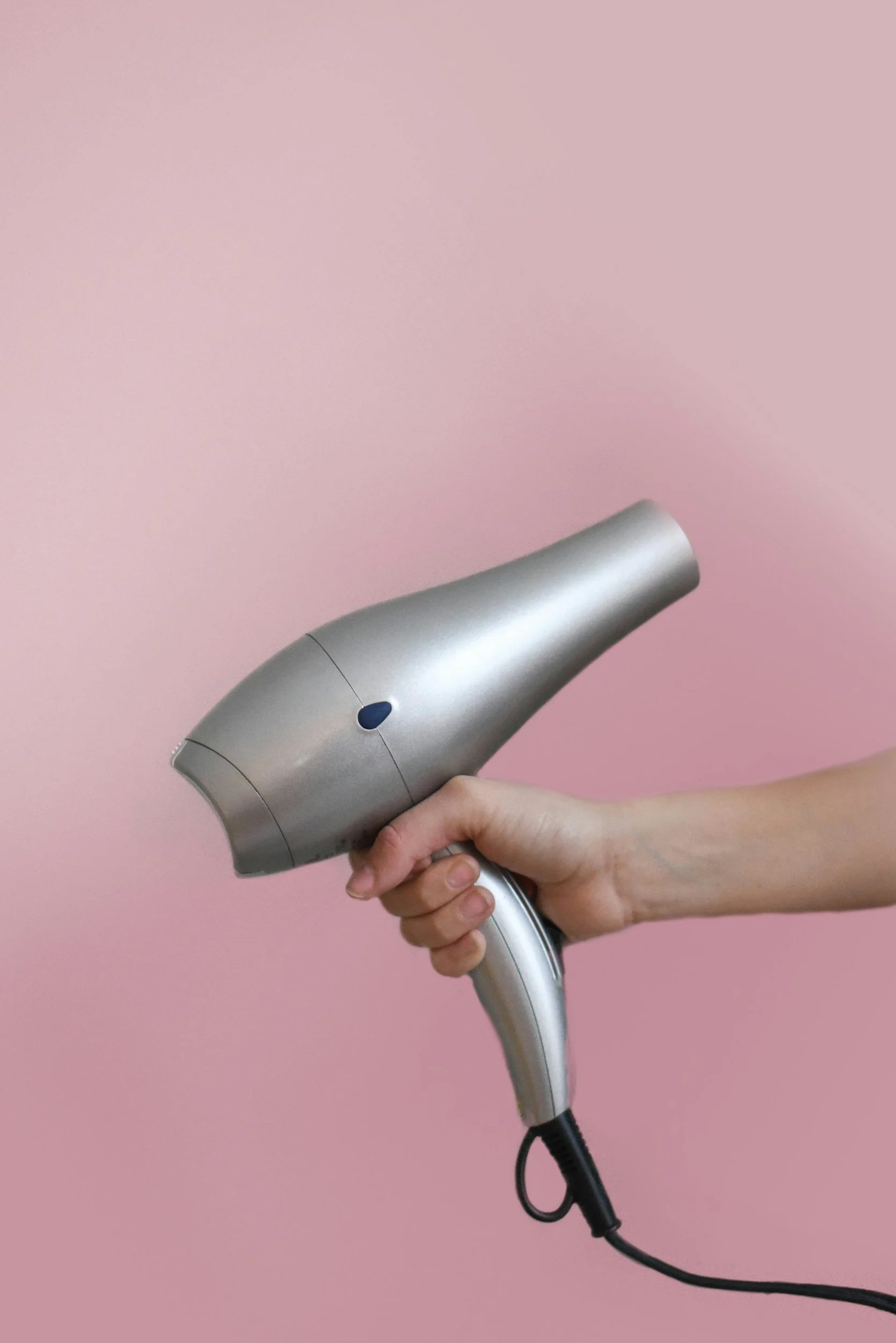 A hand holding a silver hairdryer against a pink background.