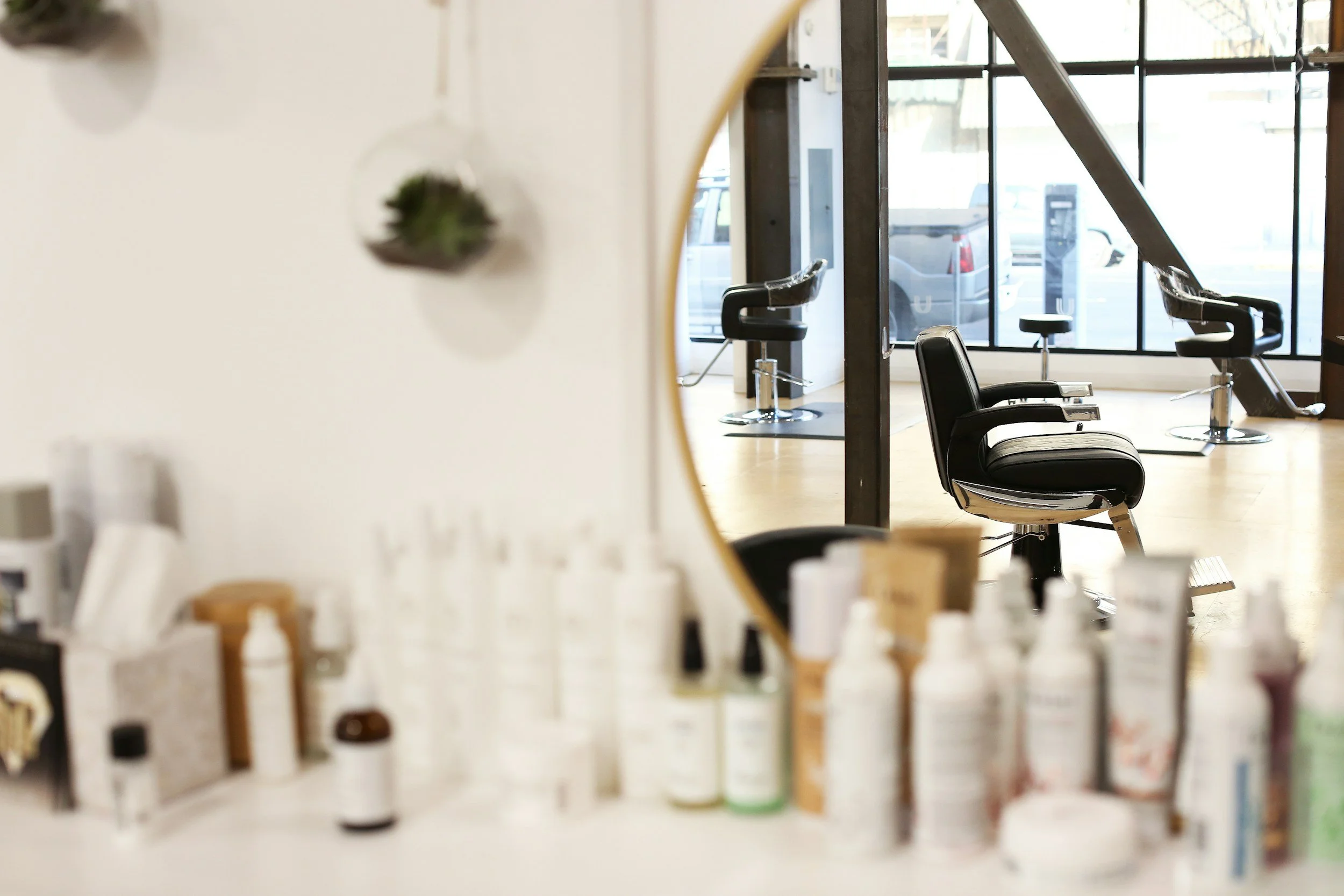 Interior view of a hair salon with empty styling chairs and large windows, seen through a blurred foreground of various hair products.